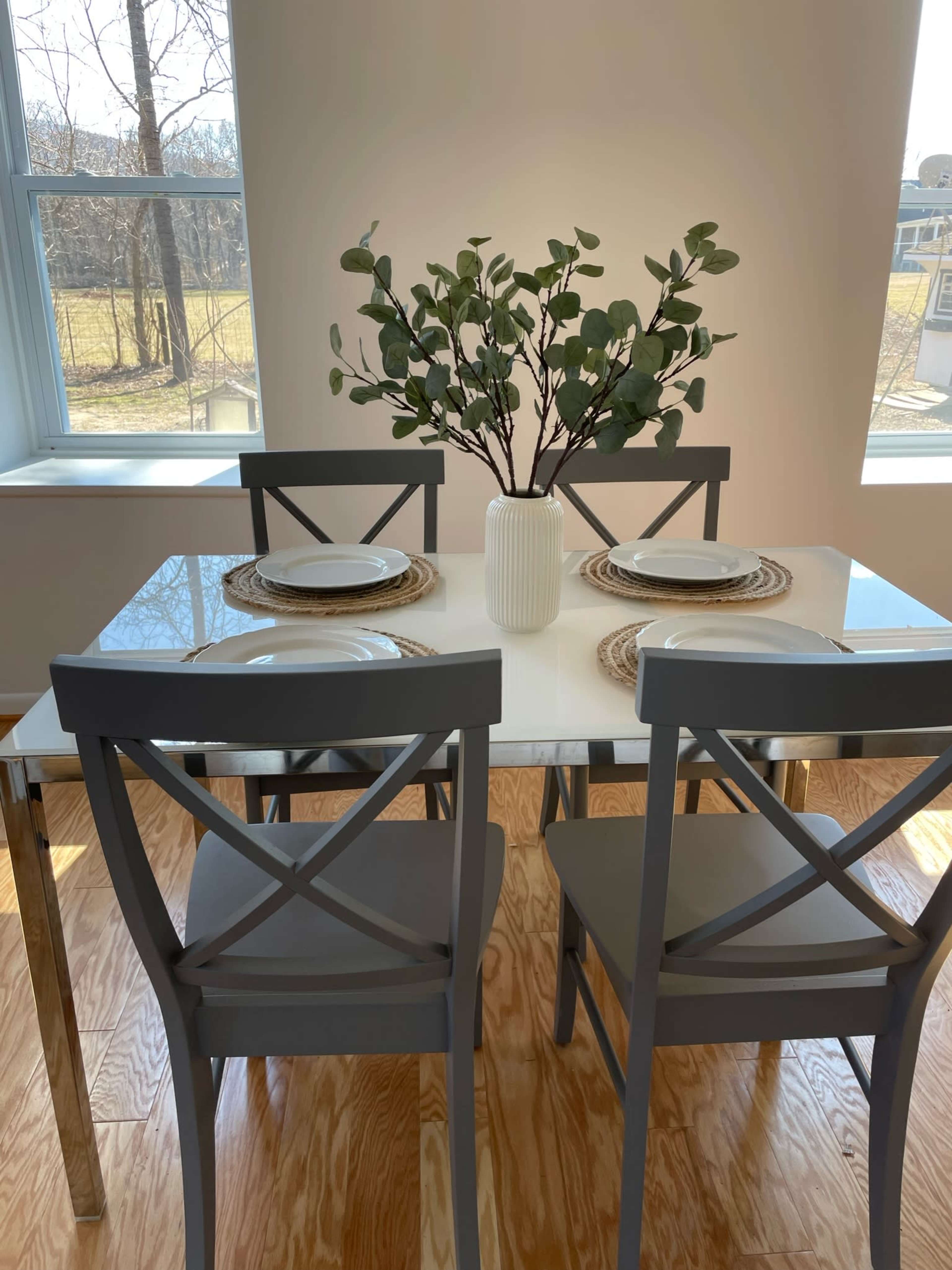 A dining table set for two, featuring plates and a vase with branches, is positioned beside large windows with natural light.