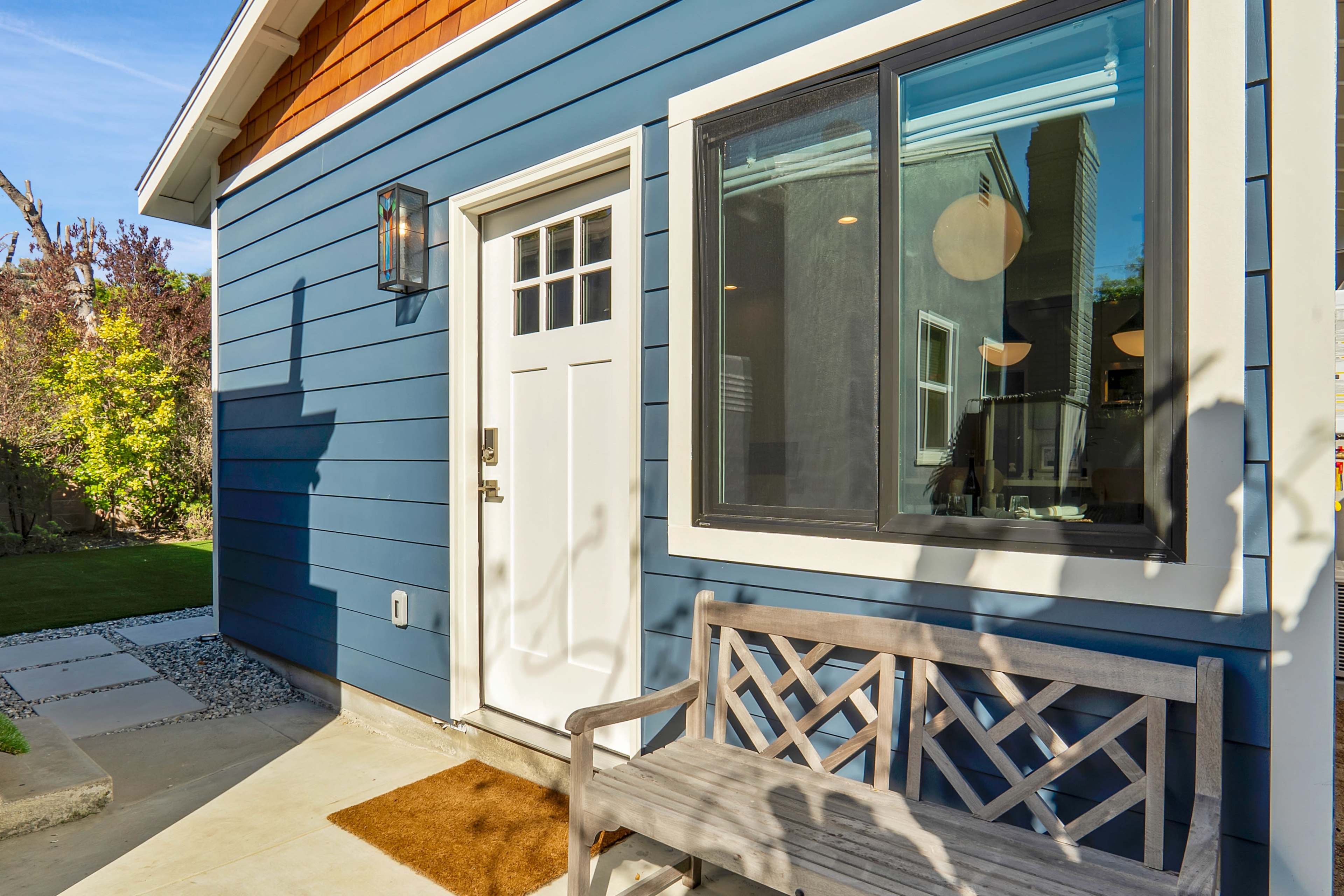 A blue house with a white front door and a wooden bench sits beside a window on a sunny day.