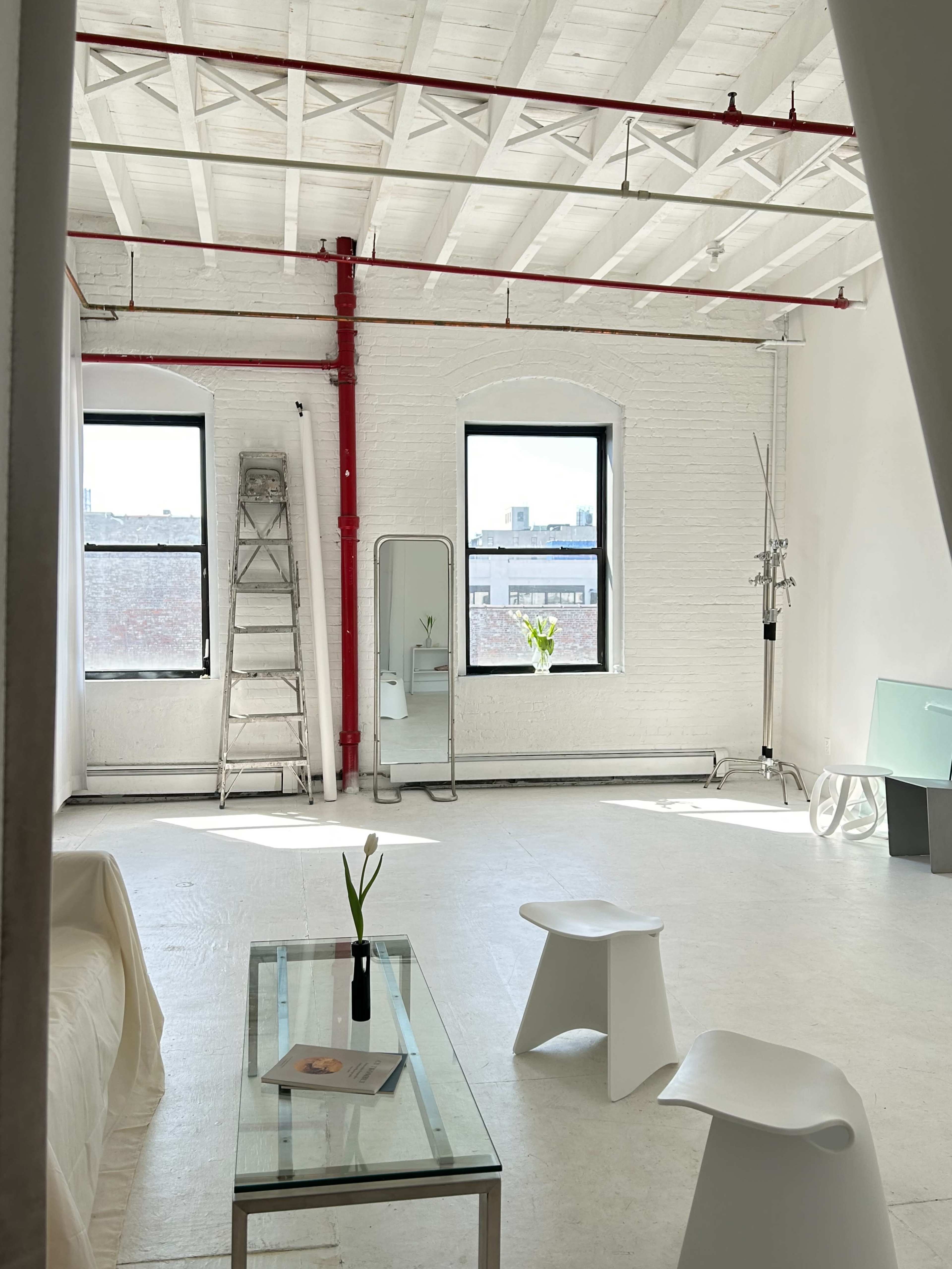 A minimalist interior space with white walls, two windows, a glass table, two white chairs, a ladder, and a potted plant.