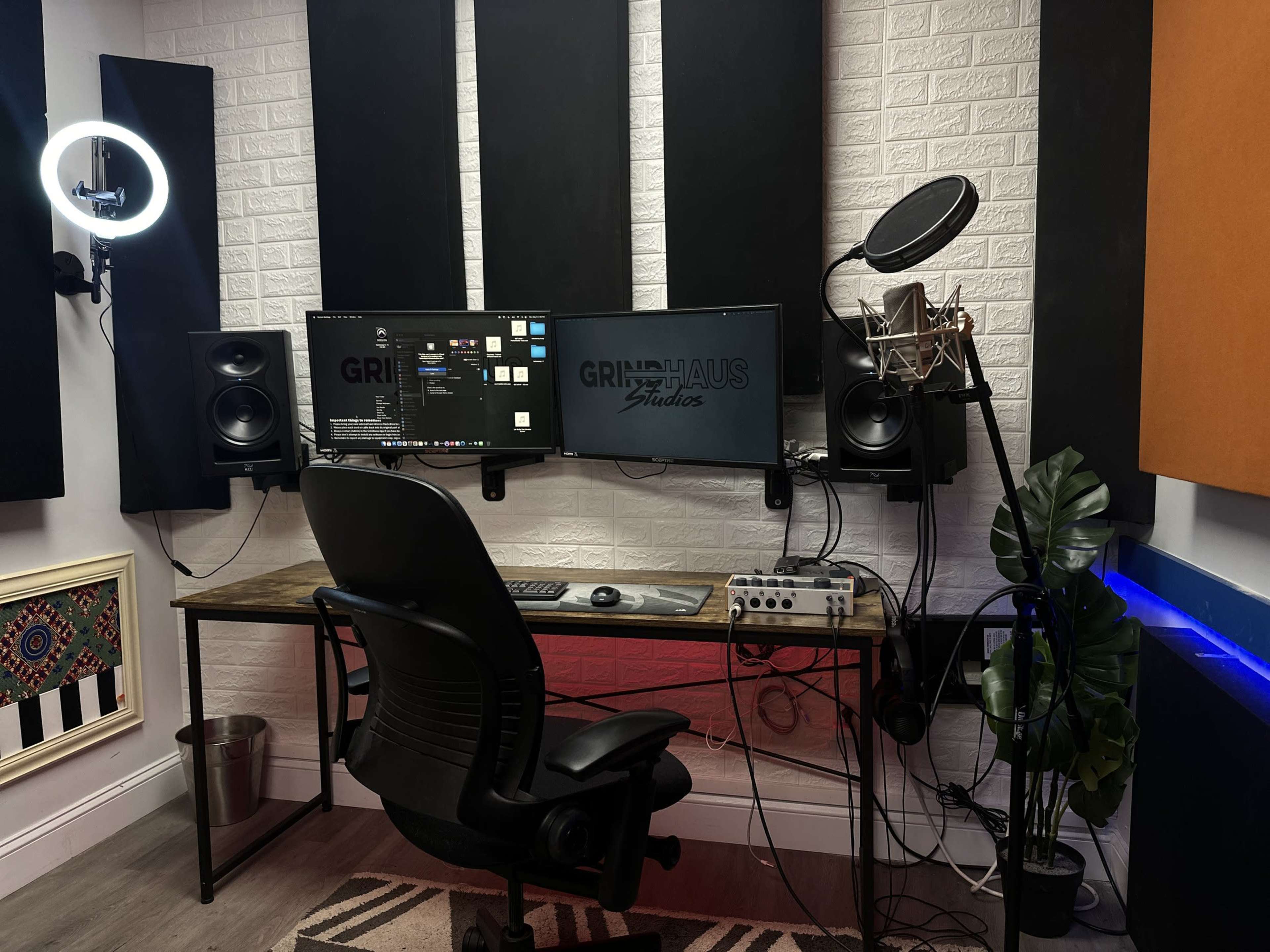 A recording studio with a desk equipped with two monitors, a microphone on a stand, a mixing interface, and soundproof panels on the walls.