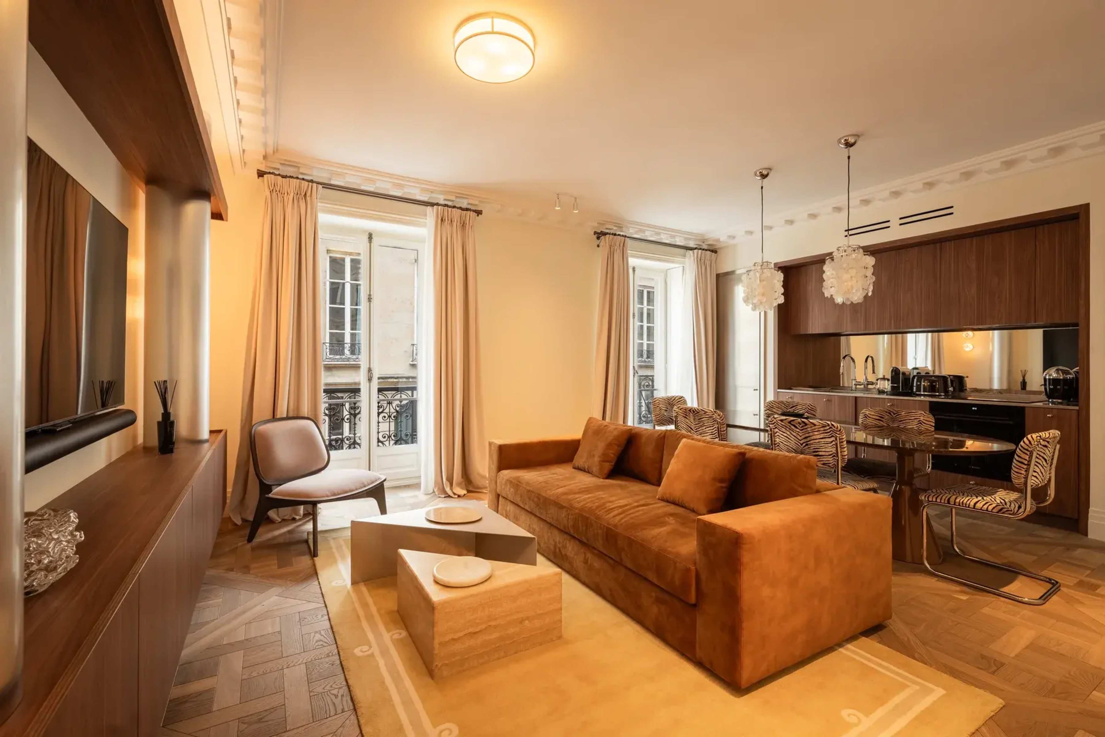 The image shows a stylish living room featuring a brown sofa, a decorative coffee table, and a dining area with elegant chairs and a modern kitchenette.