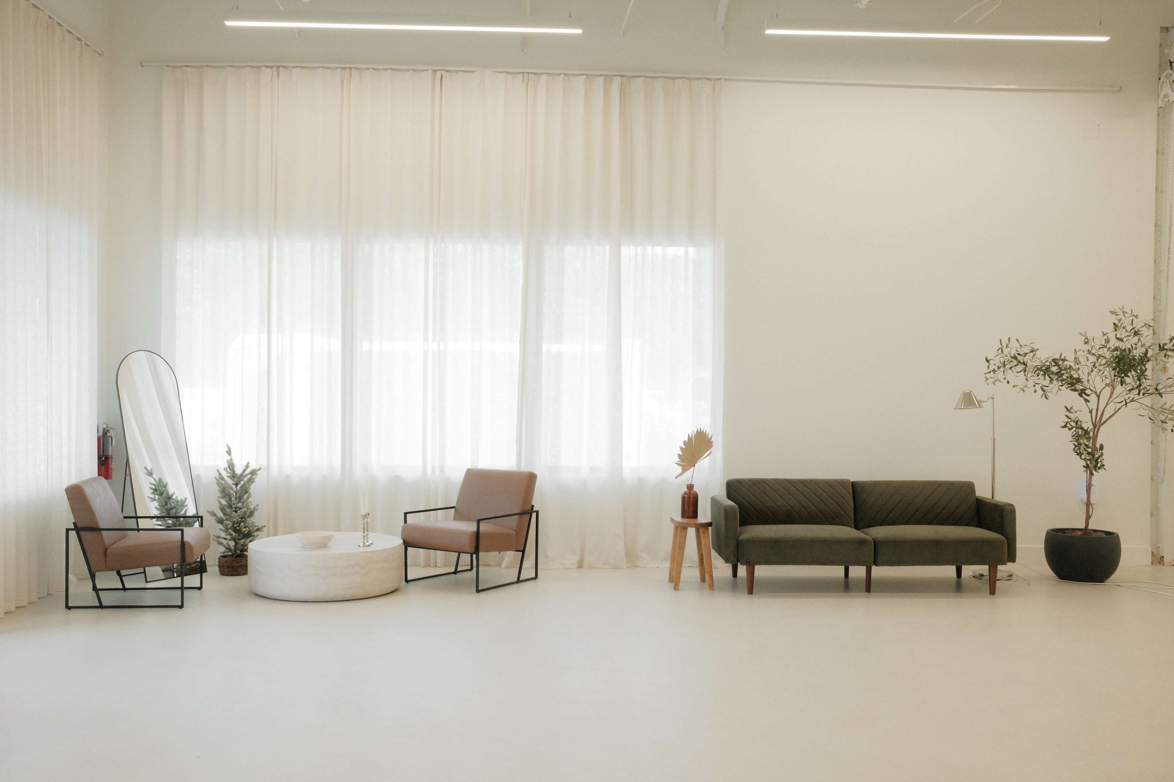 A spacious, minimalist room features a pair of chairs, a sofa, a round coffee table, a tall mirror, and potted plants near large windows with sheer curtains.