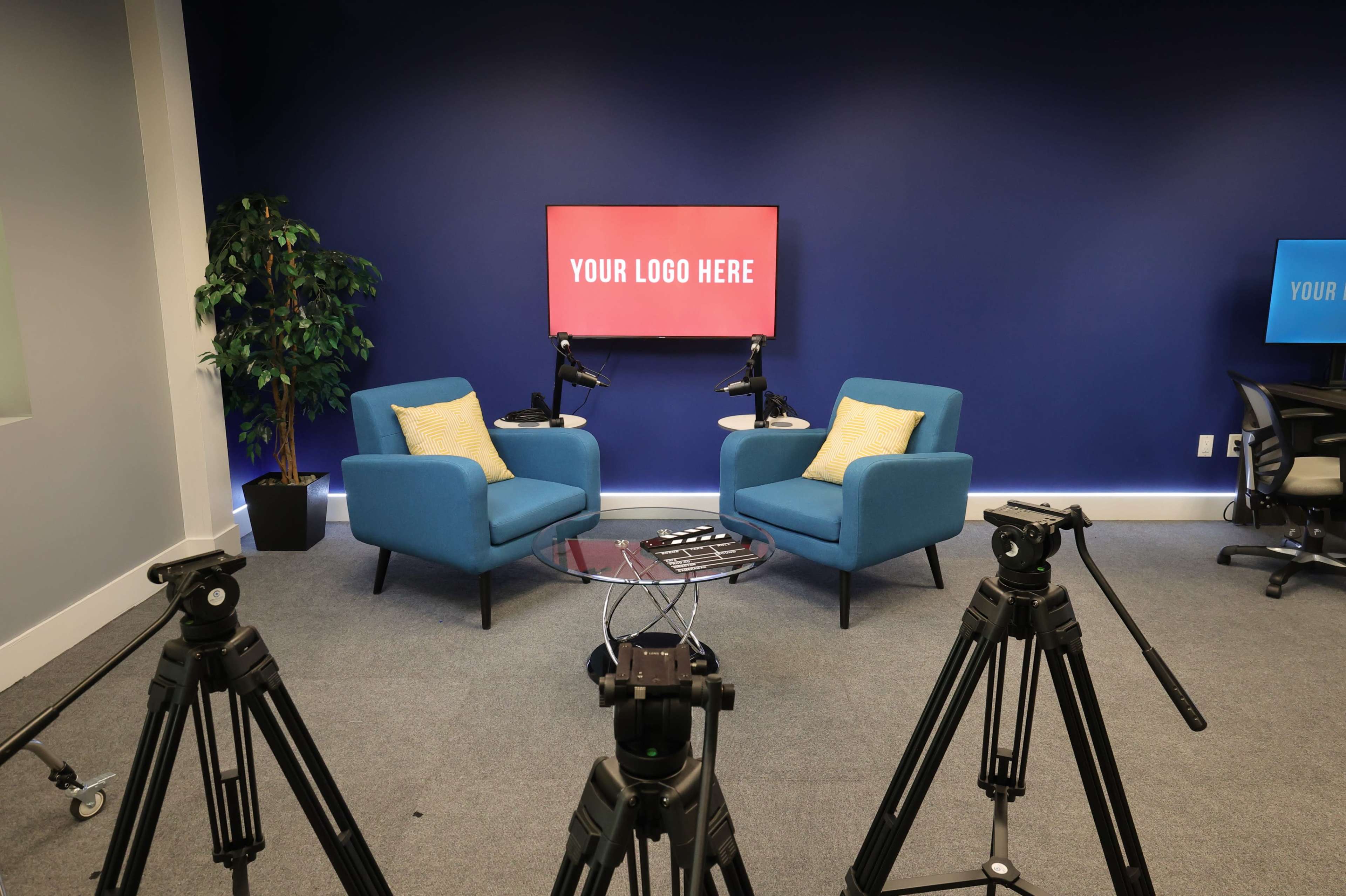 Two blue chairs with yellow cushions in a studio setting, facing a screen displaying the text "YOUR LOGO HERE," with camera tripods arranged in the foreground.