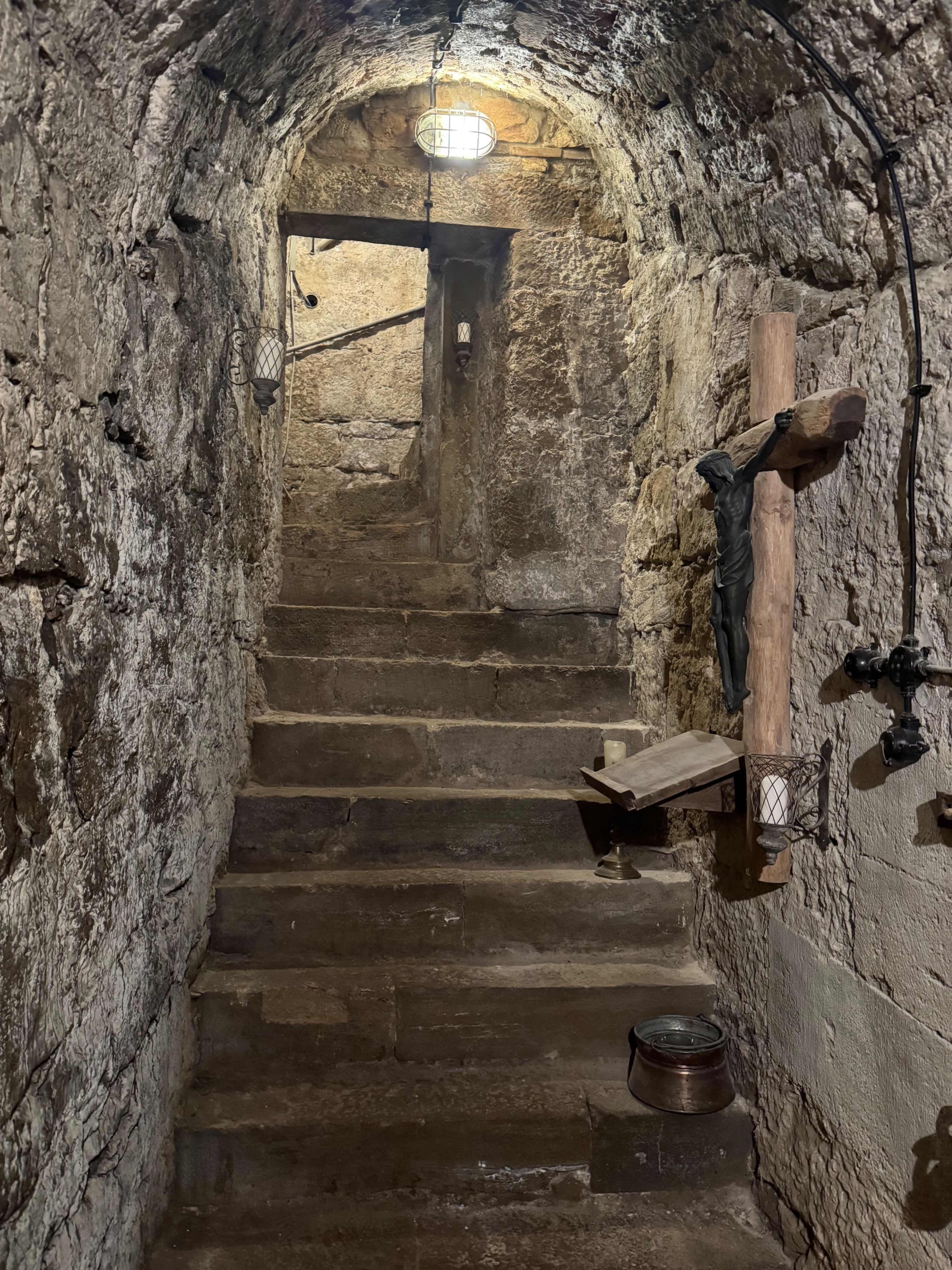 The image shows a dimly lit stone staircase leading upward, featuring rough stone walls and a wooden cross on the right side.