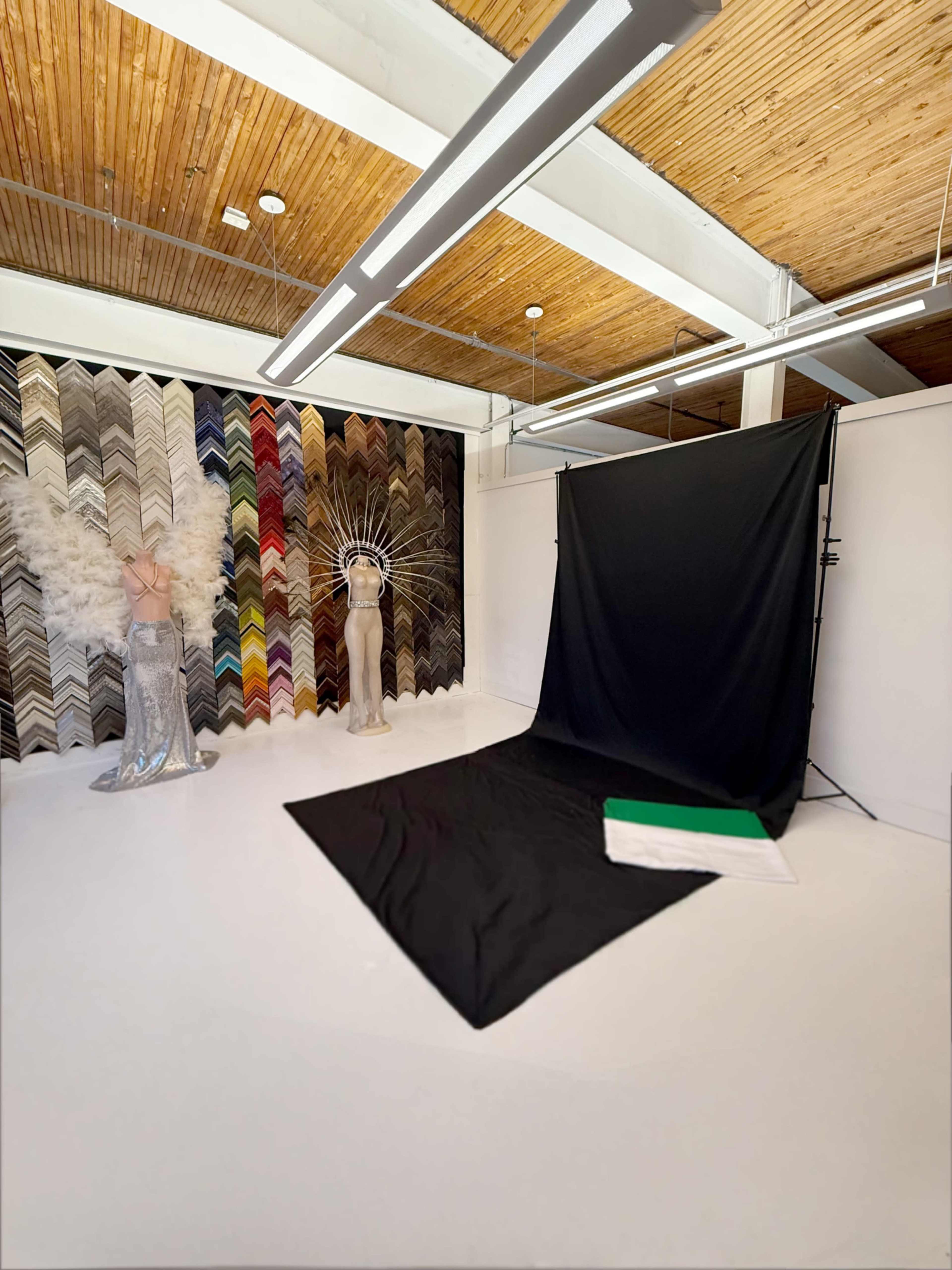 The image shows a spacious studio with a black backdrop setup, featuring decorative feathers and a colorful chevron pattern on the wall, alongside a mannequin dressed in a sparkling silver garment.
