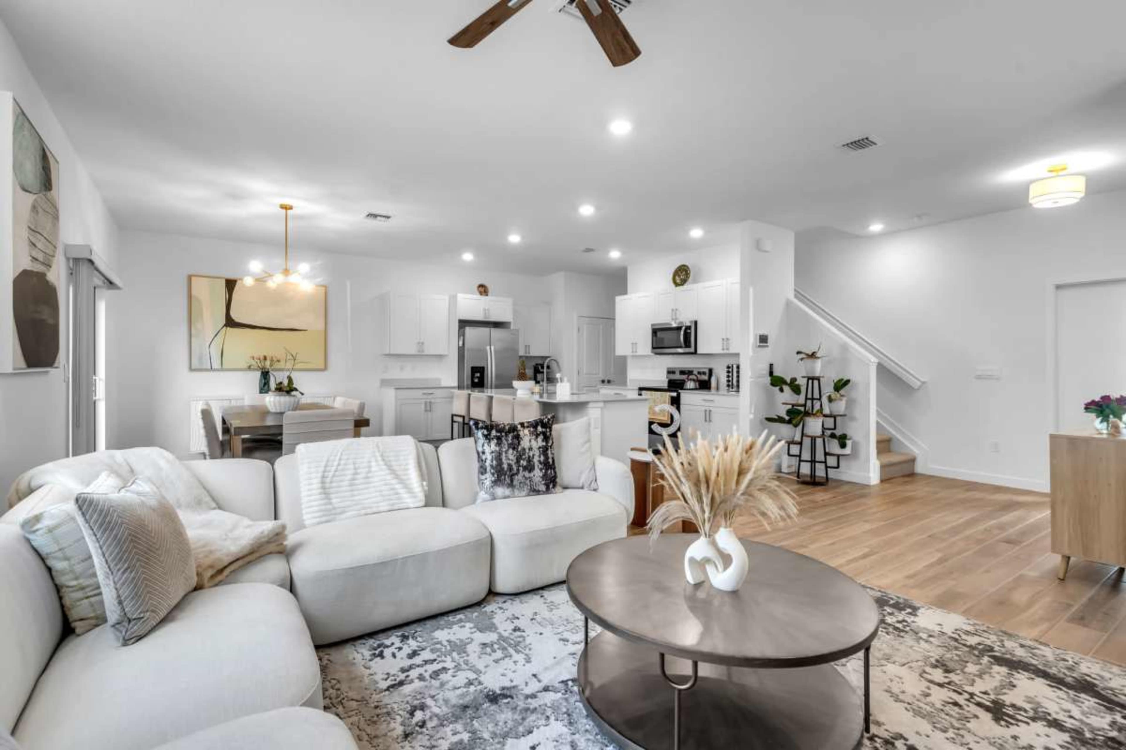 A modern, open-concept living space includes a light-colored sectional sofa, a round coffee table, a dining area, and a kitchen with white cabinetry and stainless-steel appliances.