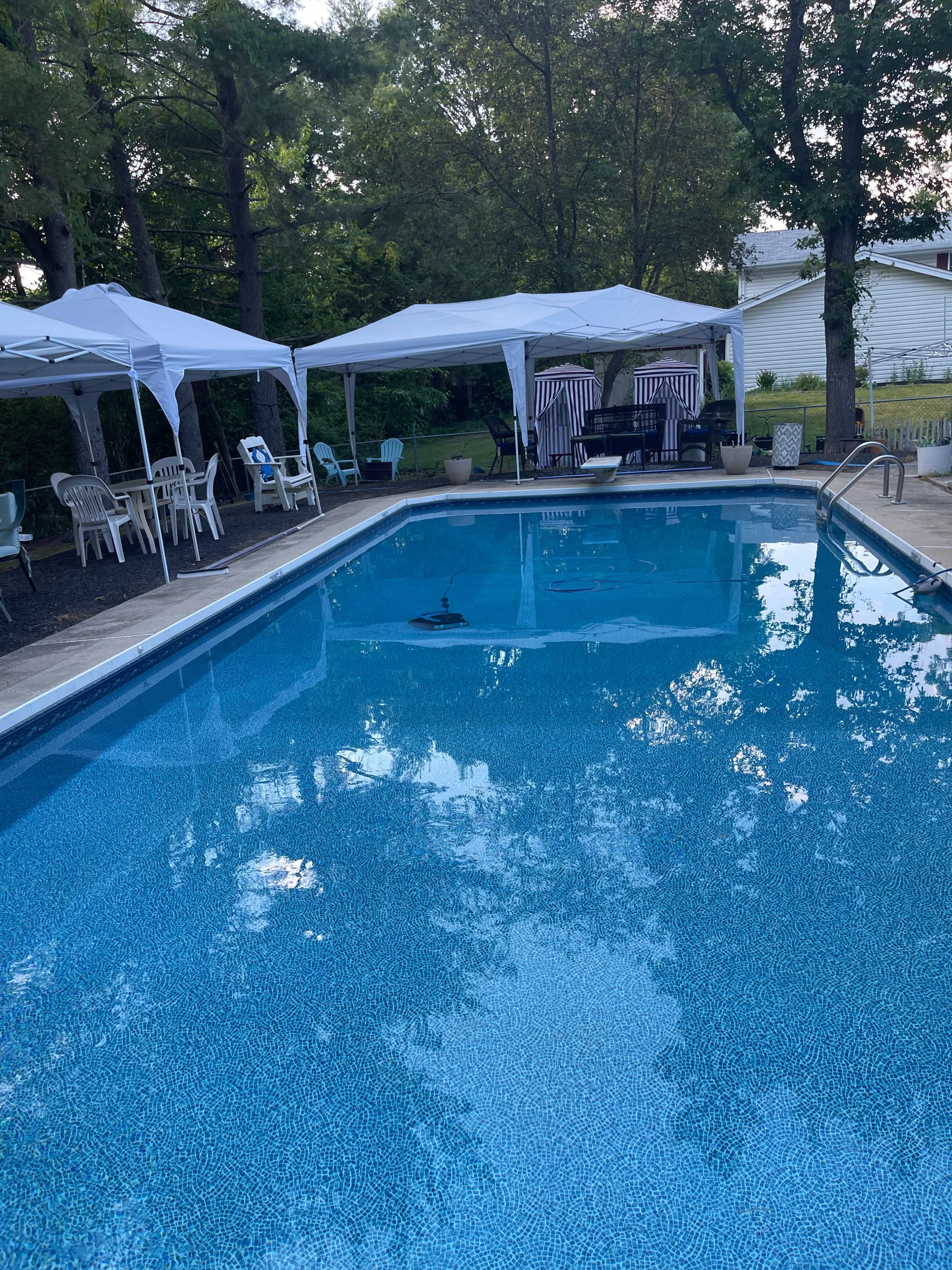 The image shows a clear swimming pool surrounded by several white canopies and a seating area, with trees and a house in the background.