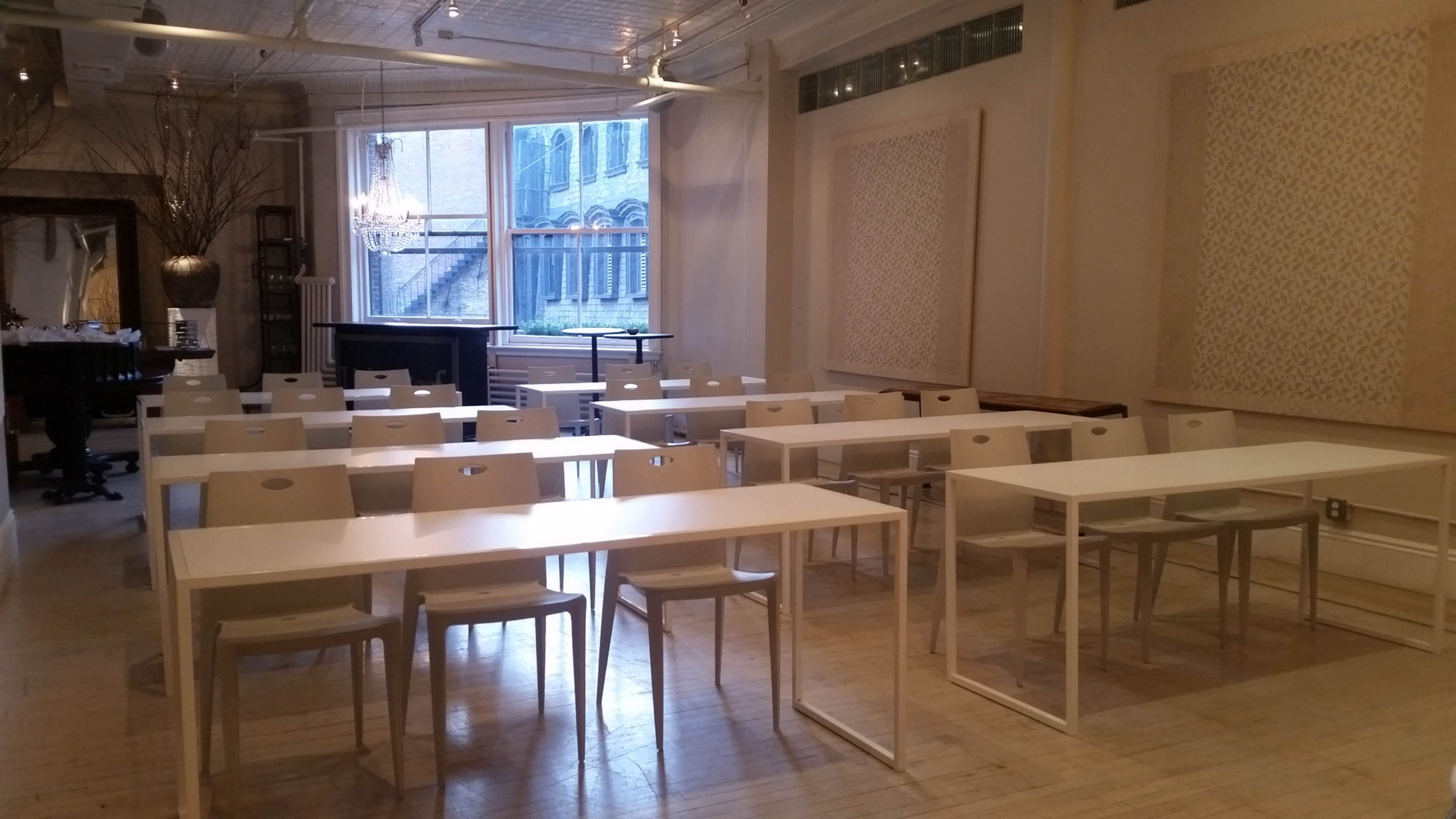 The image shows a spacious room with multiple white tables and chairs arranged in rows, featuring large windows that allow natural light to enter.