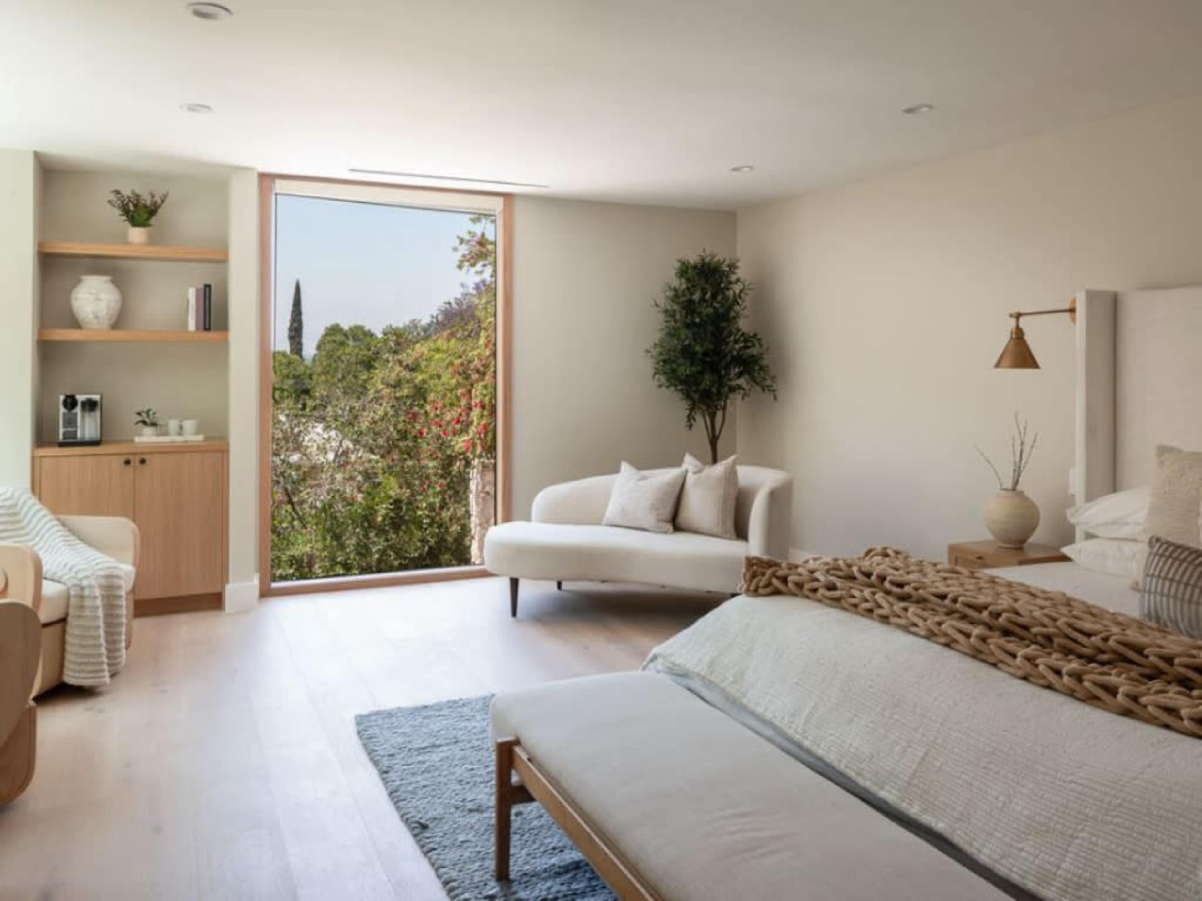 A bright bedroom features a large window overlooking greenery, with a bed, a couch, and a small shelf with decorative items.