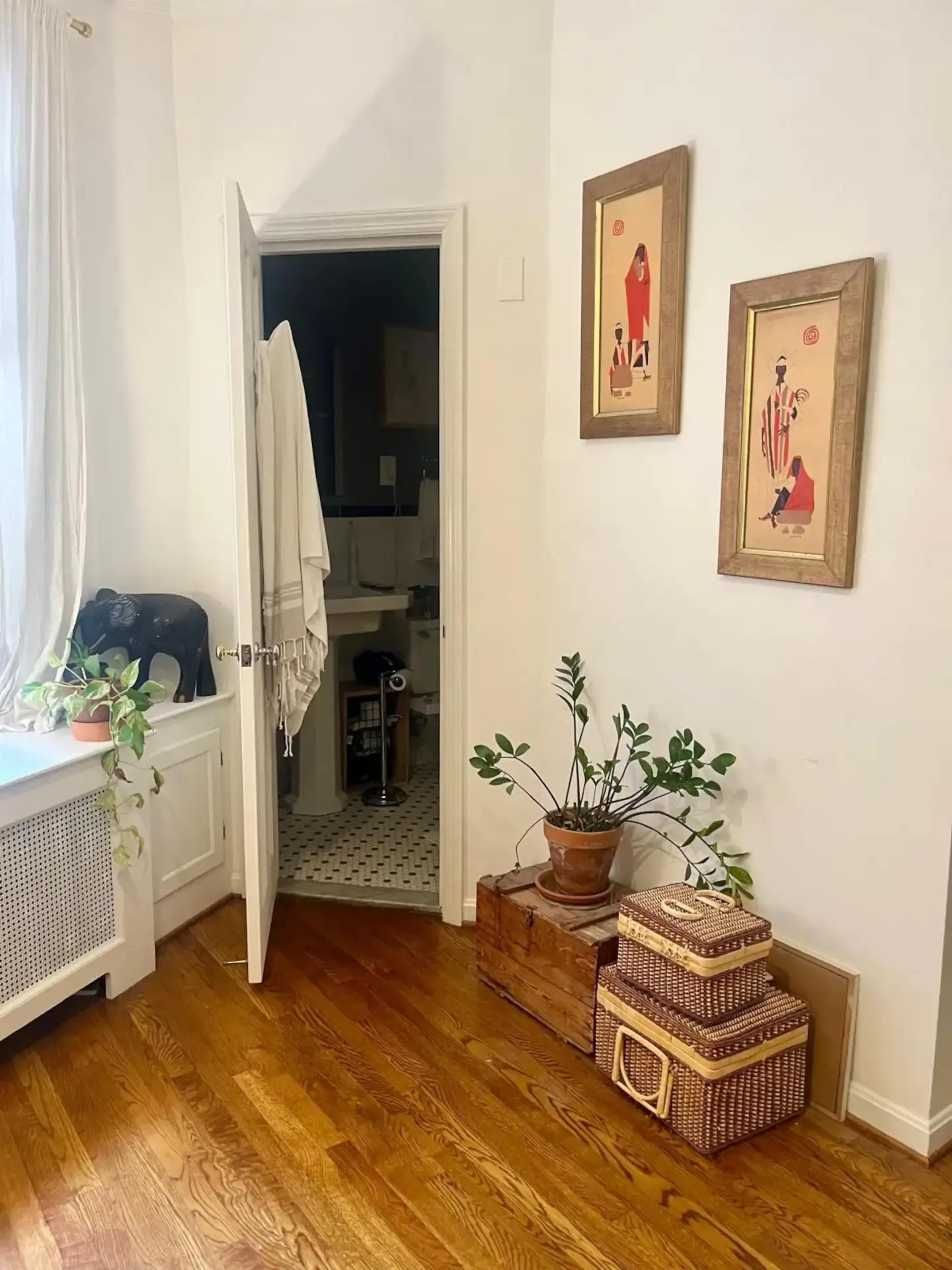 A doorway leads to a bathroom, while framed artwork, a potted plant, and decorative boxes are arranged on the floor in a bright room.
