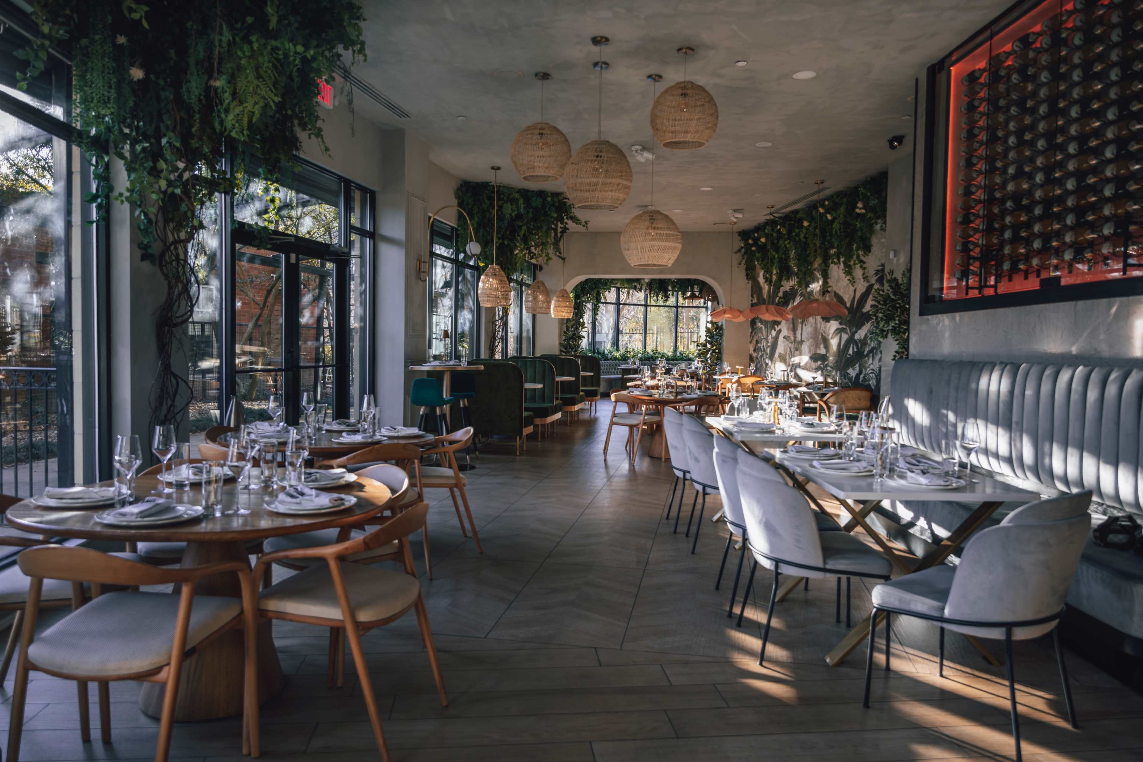 The image shows a modern restaurant interior with wooden flooring, circular and rectangular dining tables set with tableware, and large windows allowing natural light to enter the space.