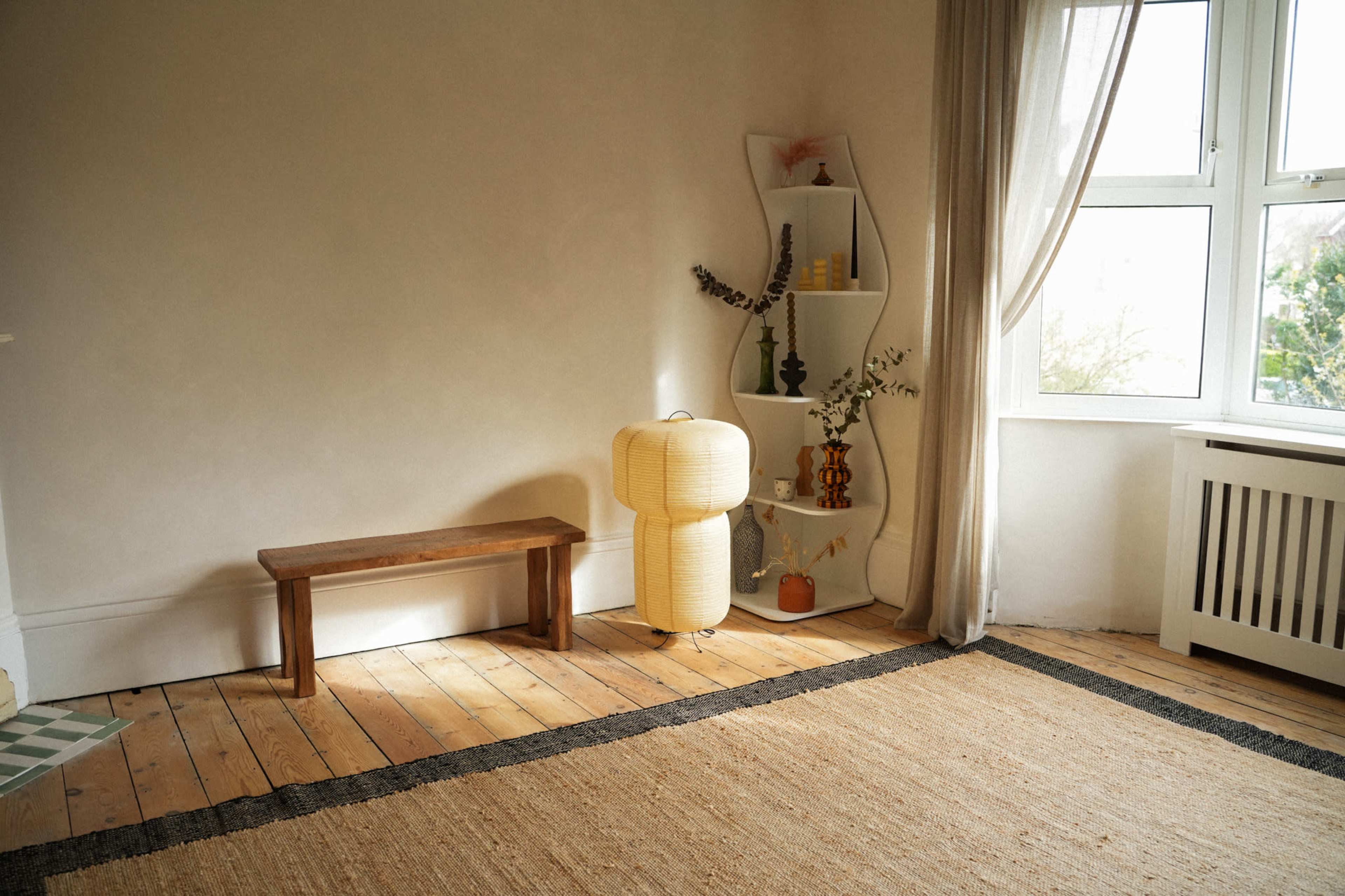 The image showcases a simple room with a wooden bench, a woven rug, and a small shelving unit displaying decorative items beside a large window.