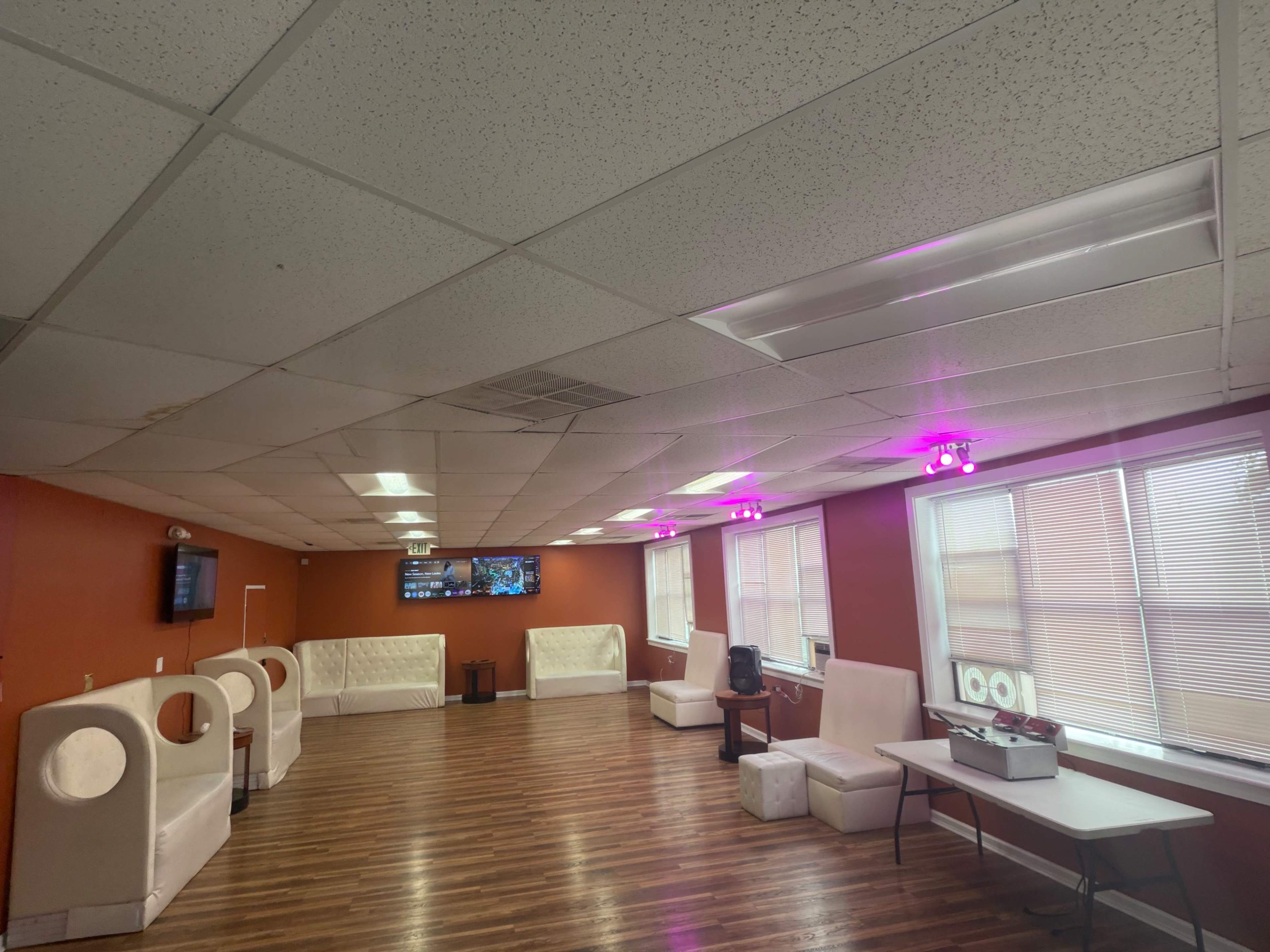 The image shows a spacious indoor lounge area with white furniture, wooden flooring, and bright lights, featuring a TV mounted on the wall.