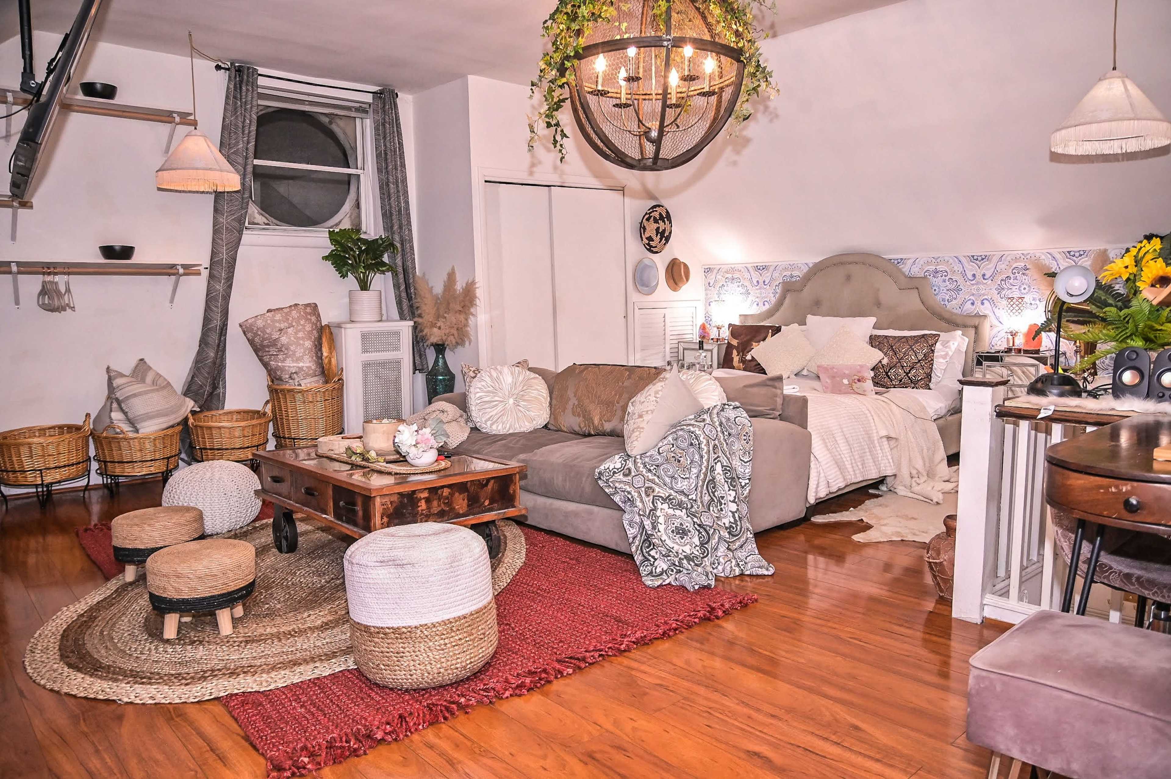The image depicts a cozy bohemian-style room featuring a large sofa, a wooden coffee table, various decorative pillows, and woven baskets, all arranged on hardwood flooring with a prominent rug.
