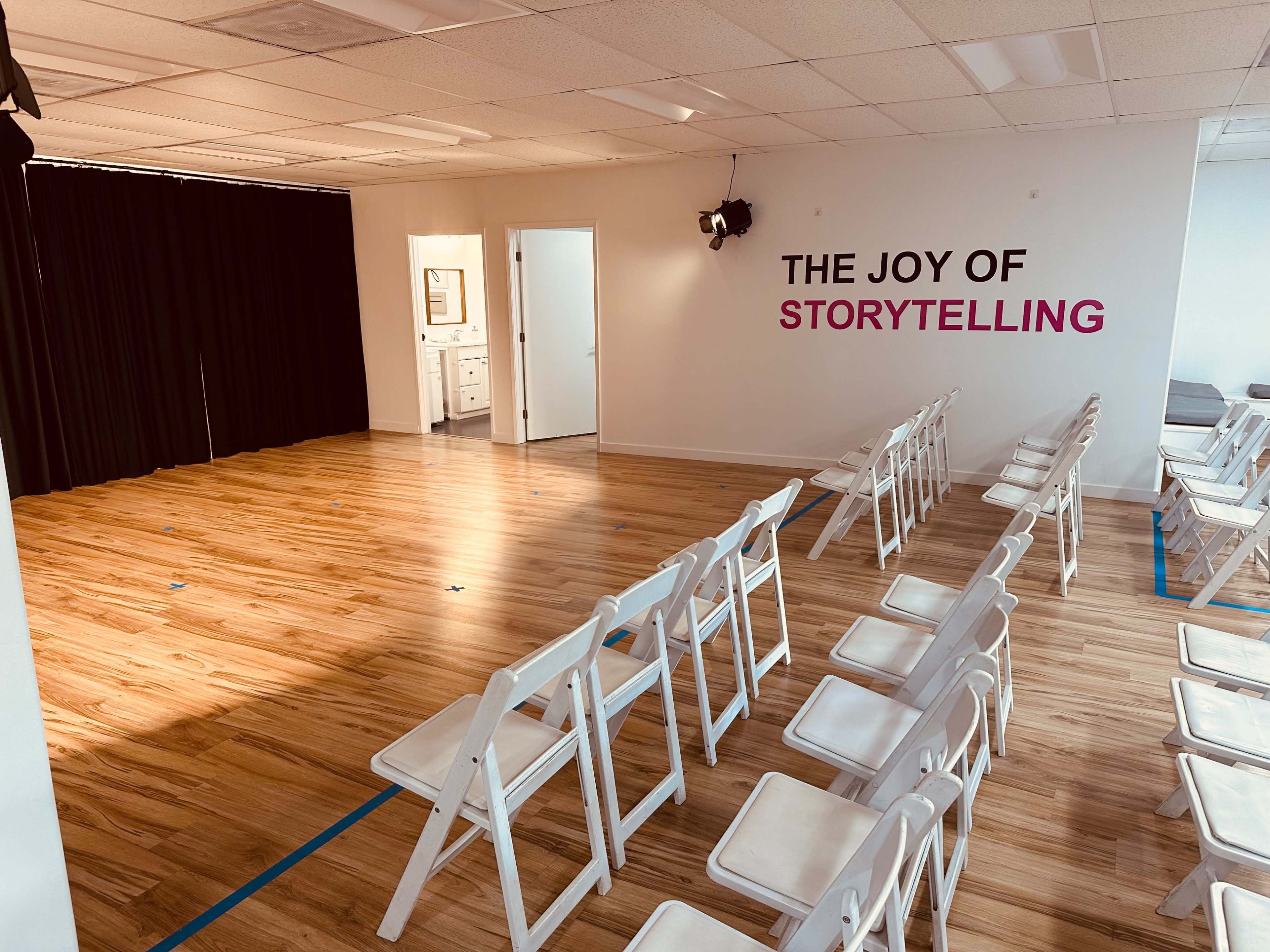 A spacious room with wooden flooring features rows of white folding chairs facing a black curtain and a wall decorated with the phrase "THE JOY OF STORYTELLING."
