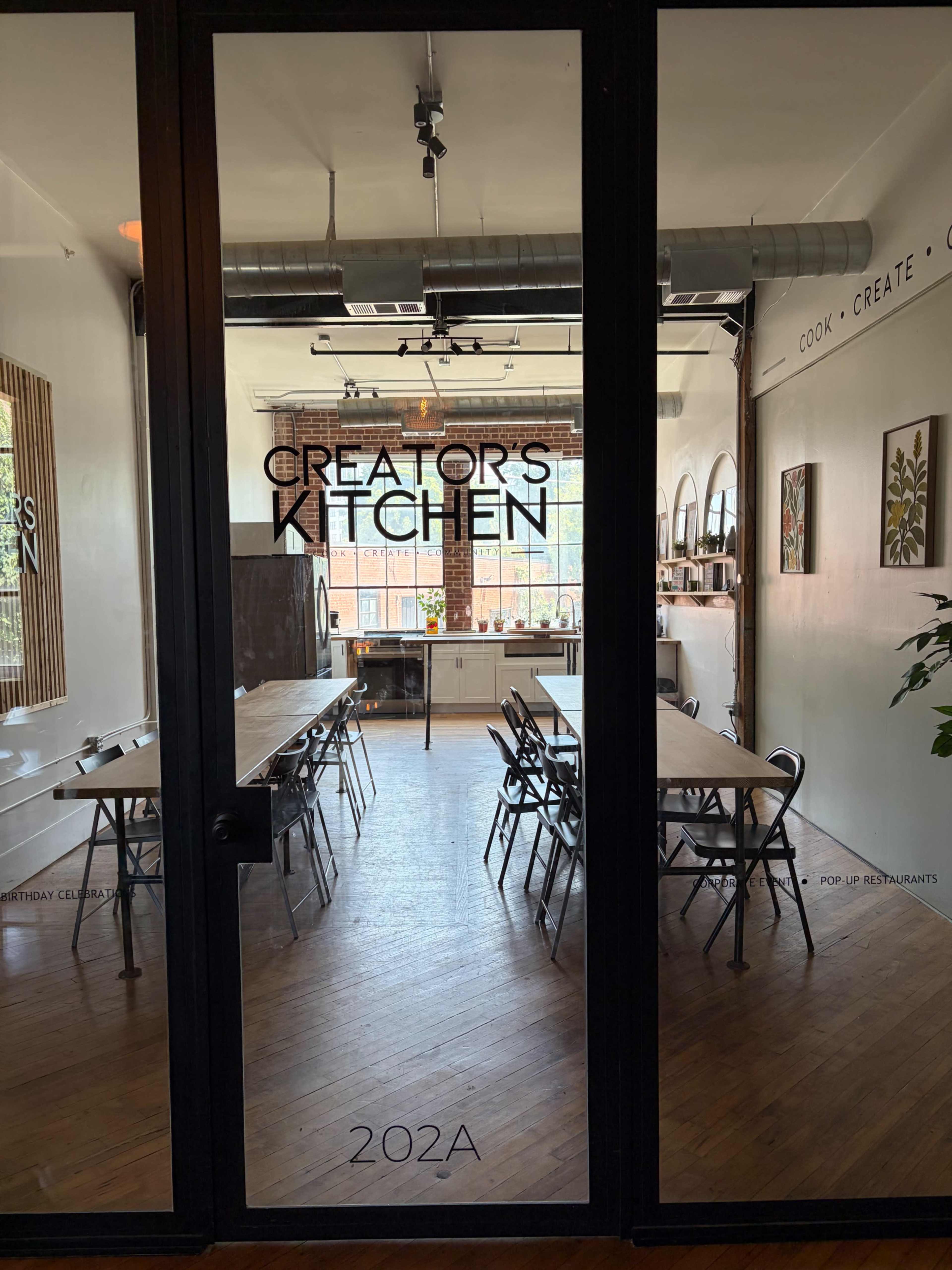The image shows the entrance to a brightly lit kitchen space, featuring long tables and chairs arranged for cooking activities.
