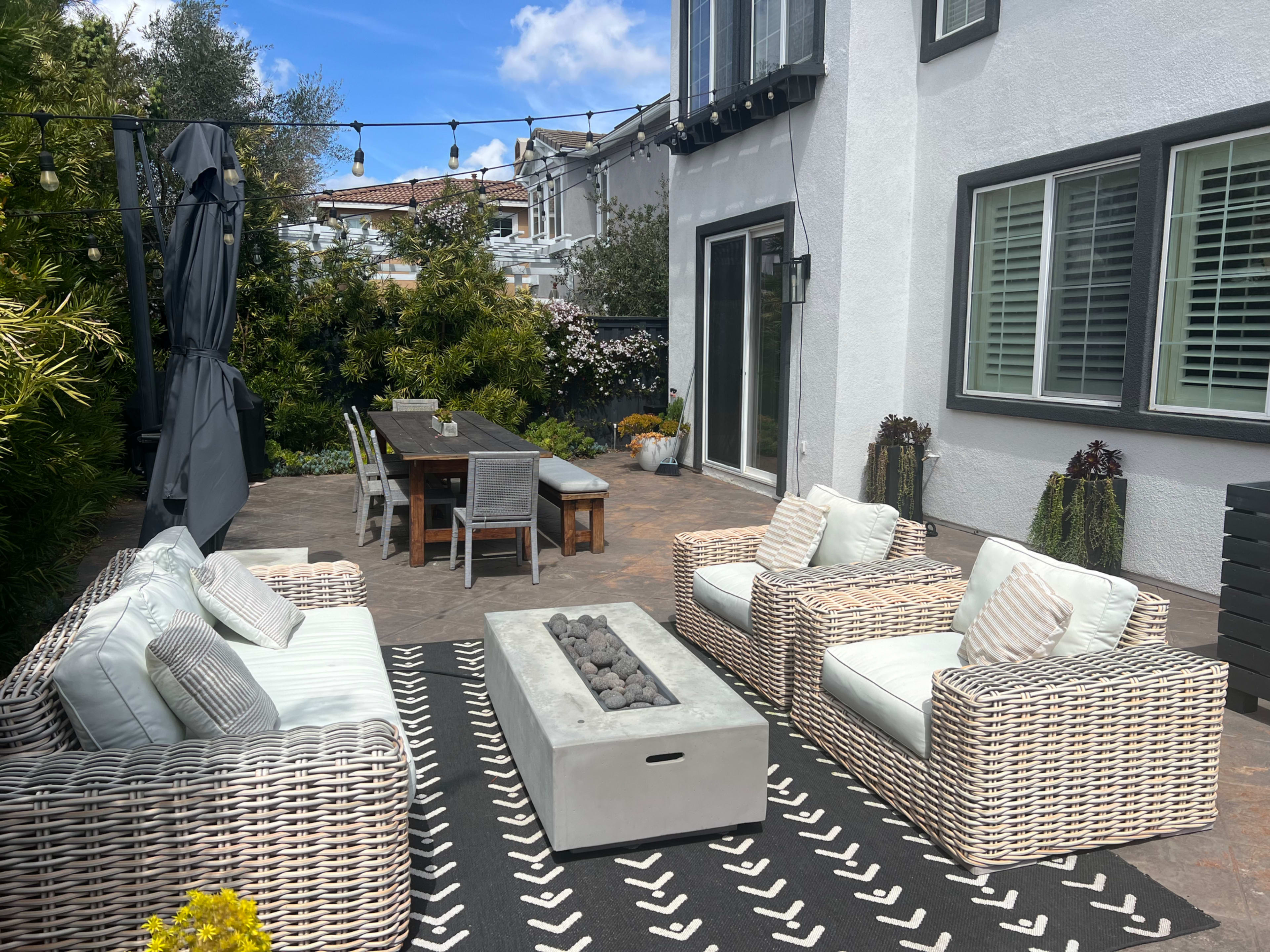 A patio area features woven furniture, a fire pit, a long dining table, and greenery, under a blue sky with string lights overhead.