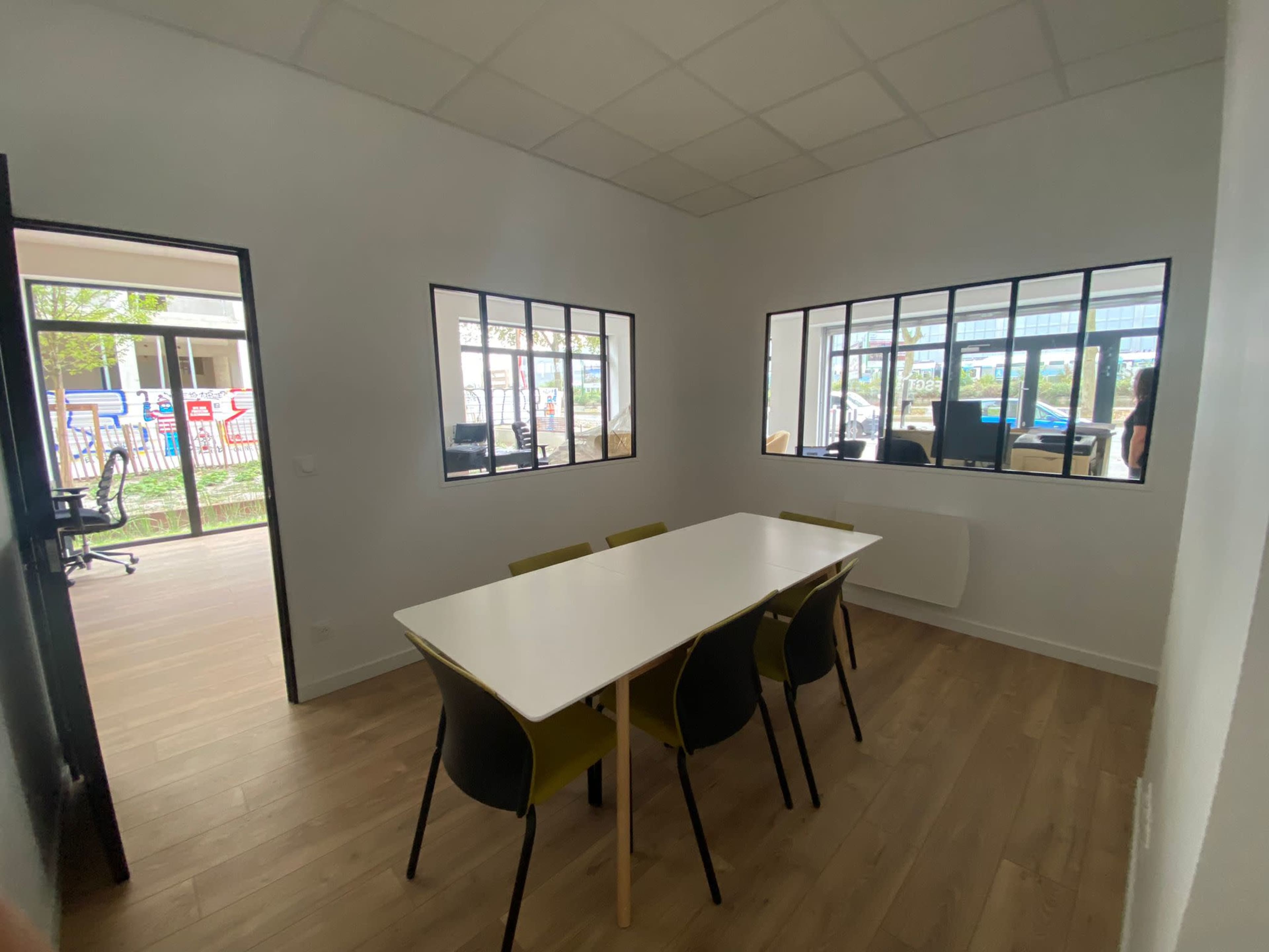 A brightly lit meeting room features a white table surrounded by four green chairs, with large windows overlooking a courtyard.