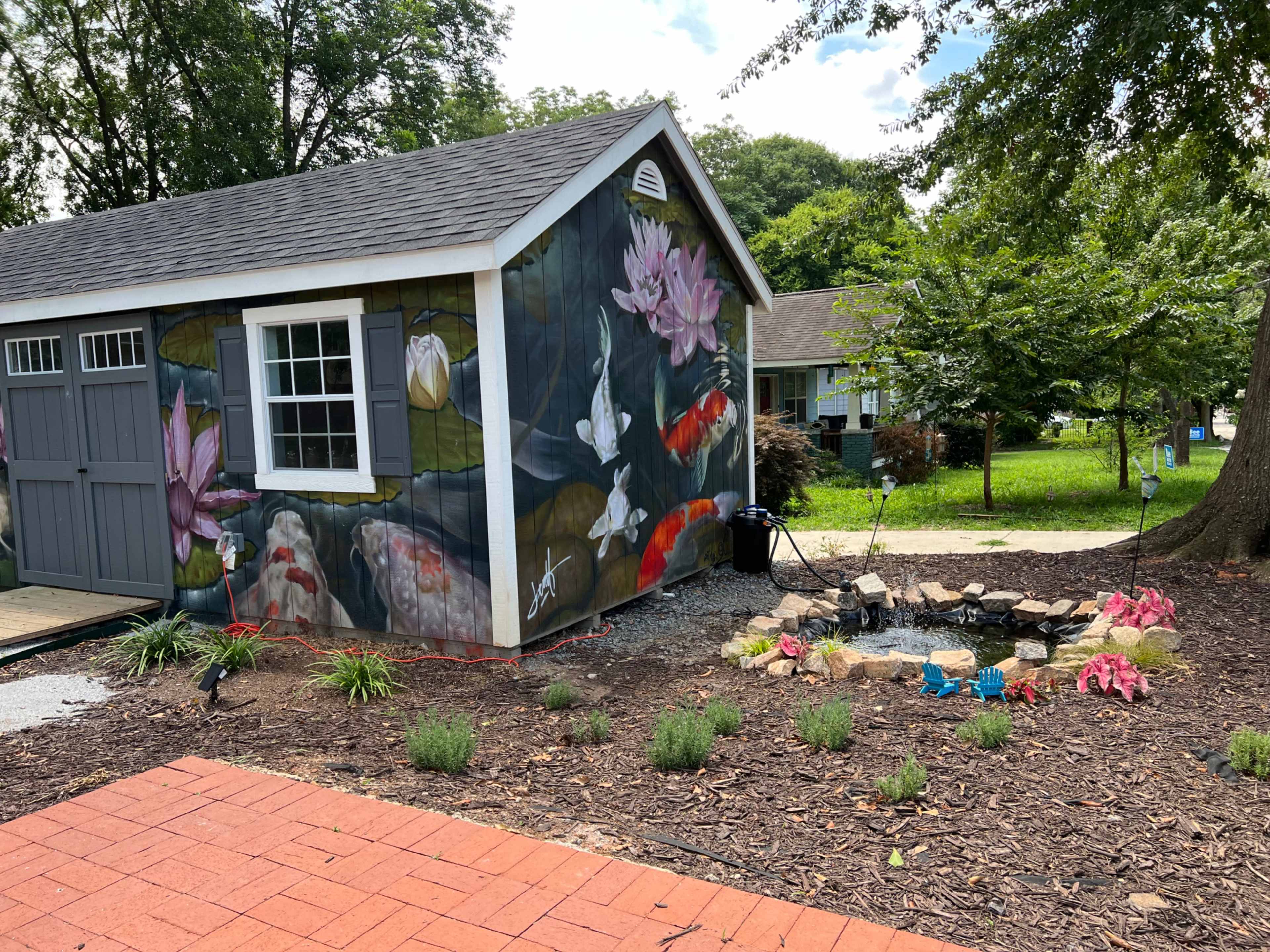 A shed features a mural of koi fish and water lilies, surrounded by a landscaped garden with flowers and stones.