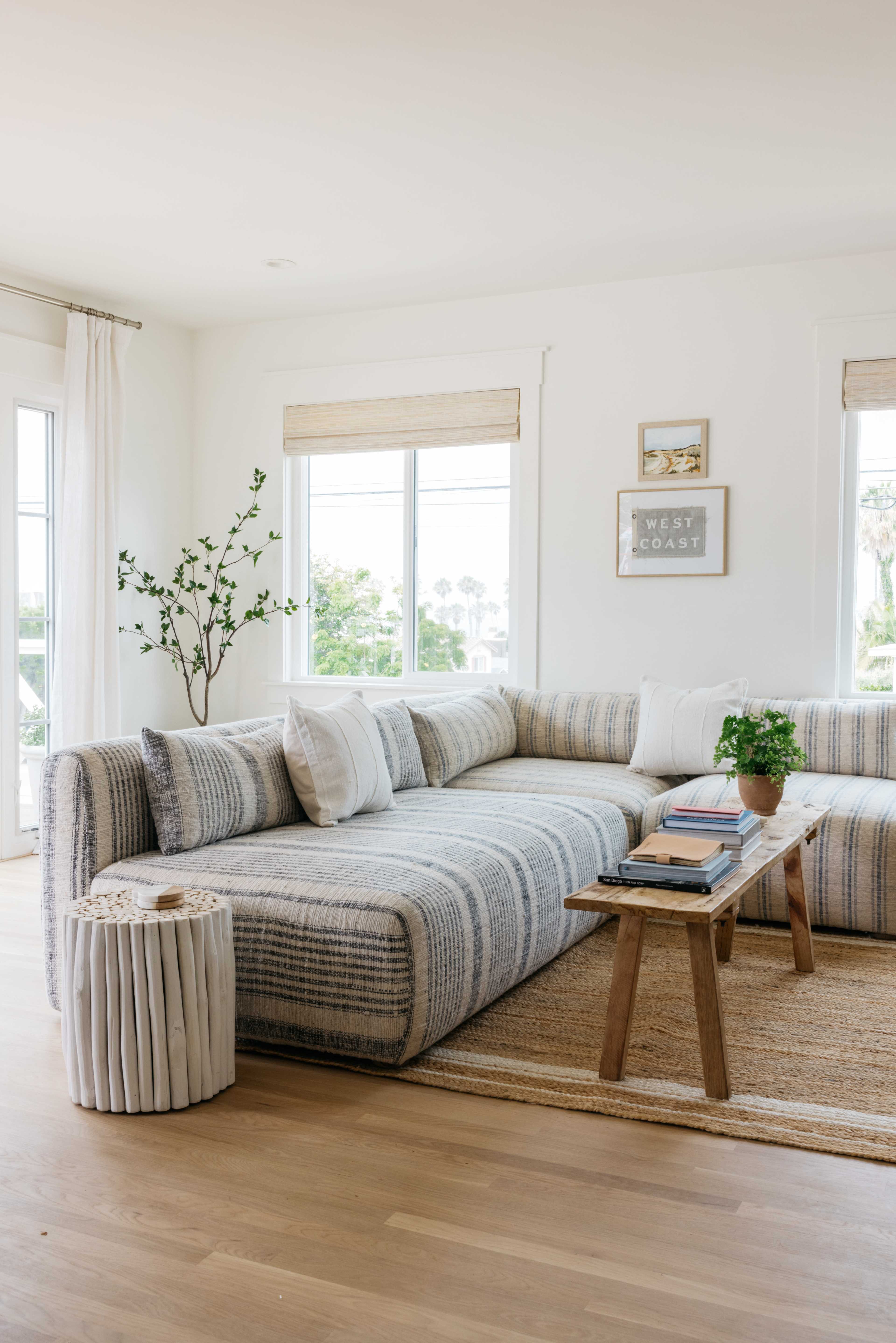 A spacious living room features a light-colored sectional sofa, a wooden coffee table, and natural light filtering through large windows.