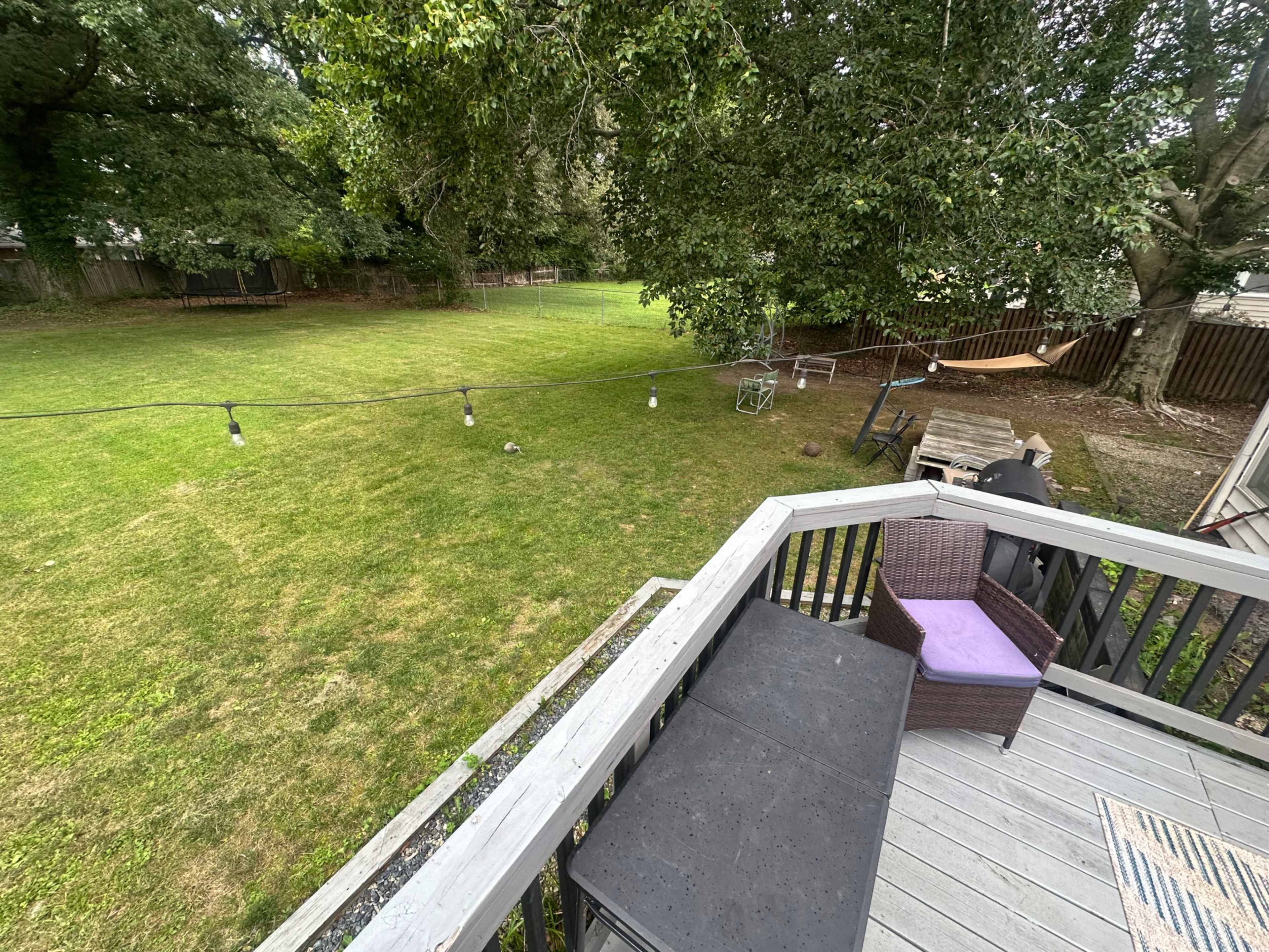 A view from a deck shows a spacious backyard with grass, a hammock, string lights, and some trees.