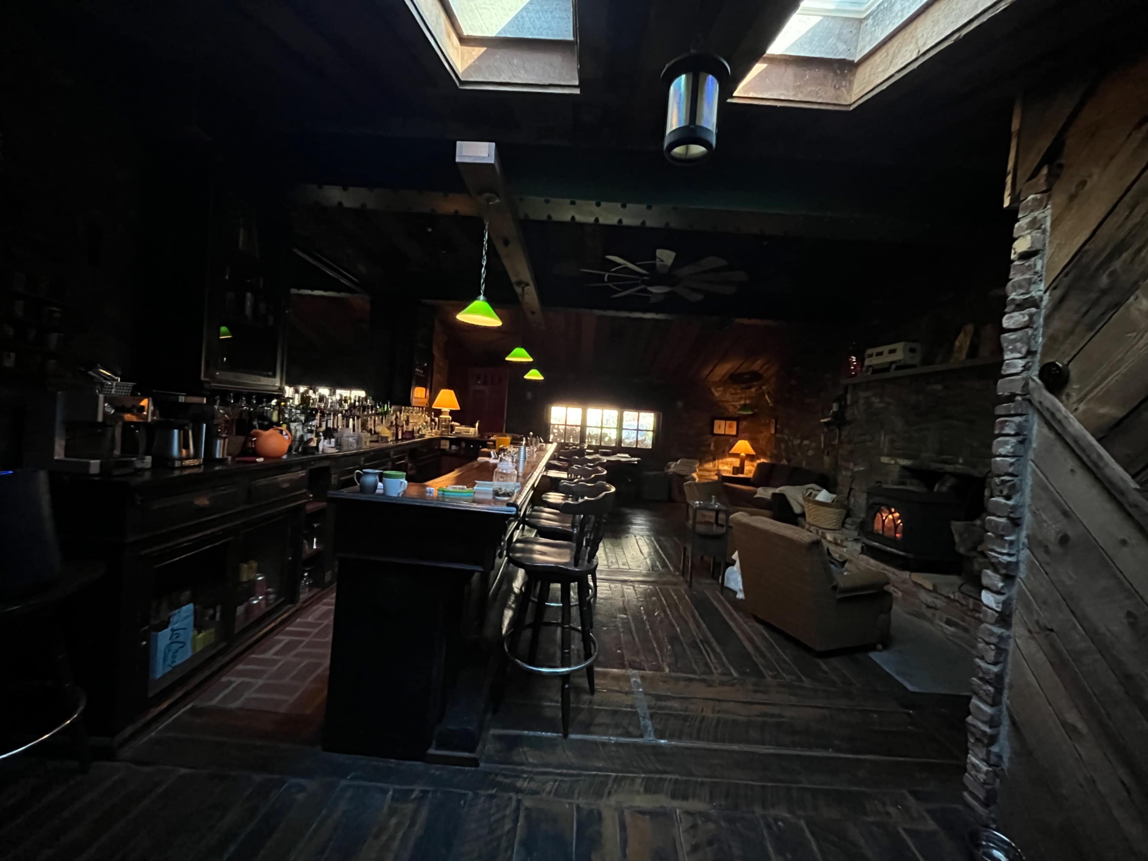 A dimly lit bar area with a wooden counter, bar stools, and a cozy seating area near a fireplace, featuring several windows and a skylight above.