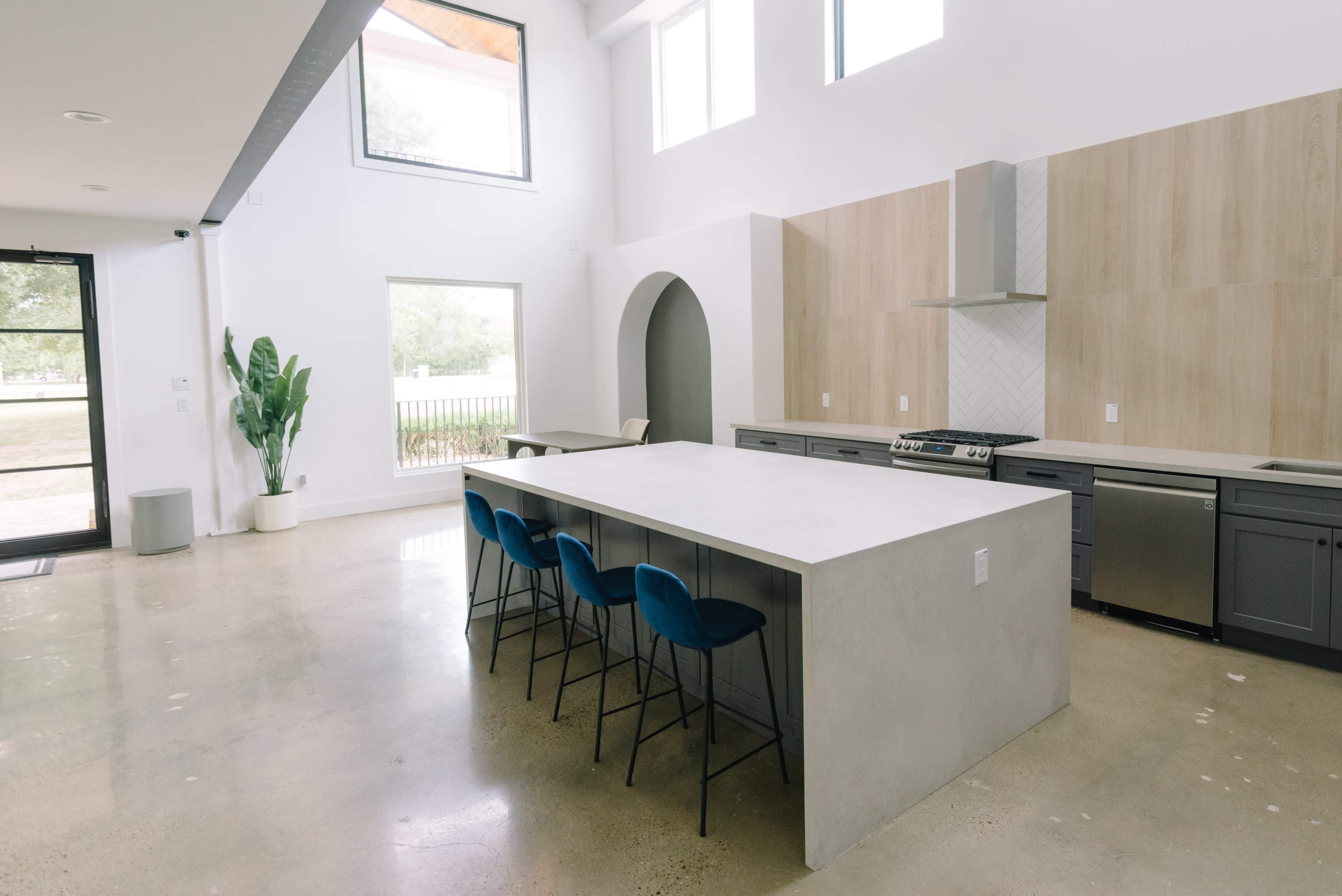 A modern kitchen features a large concrete island with four blue stools, sleek cabinetry, and ample natural light from large windows.
