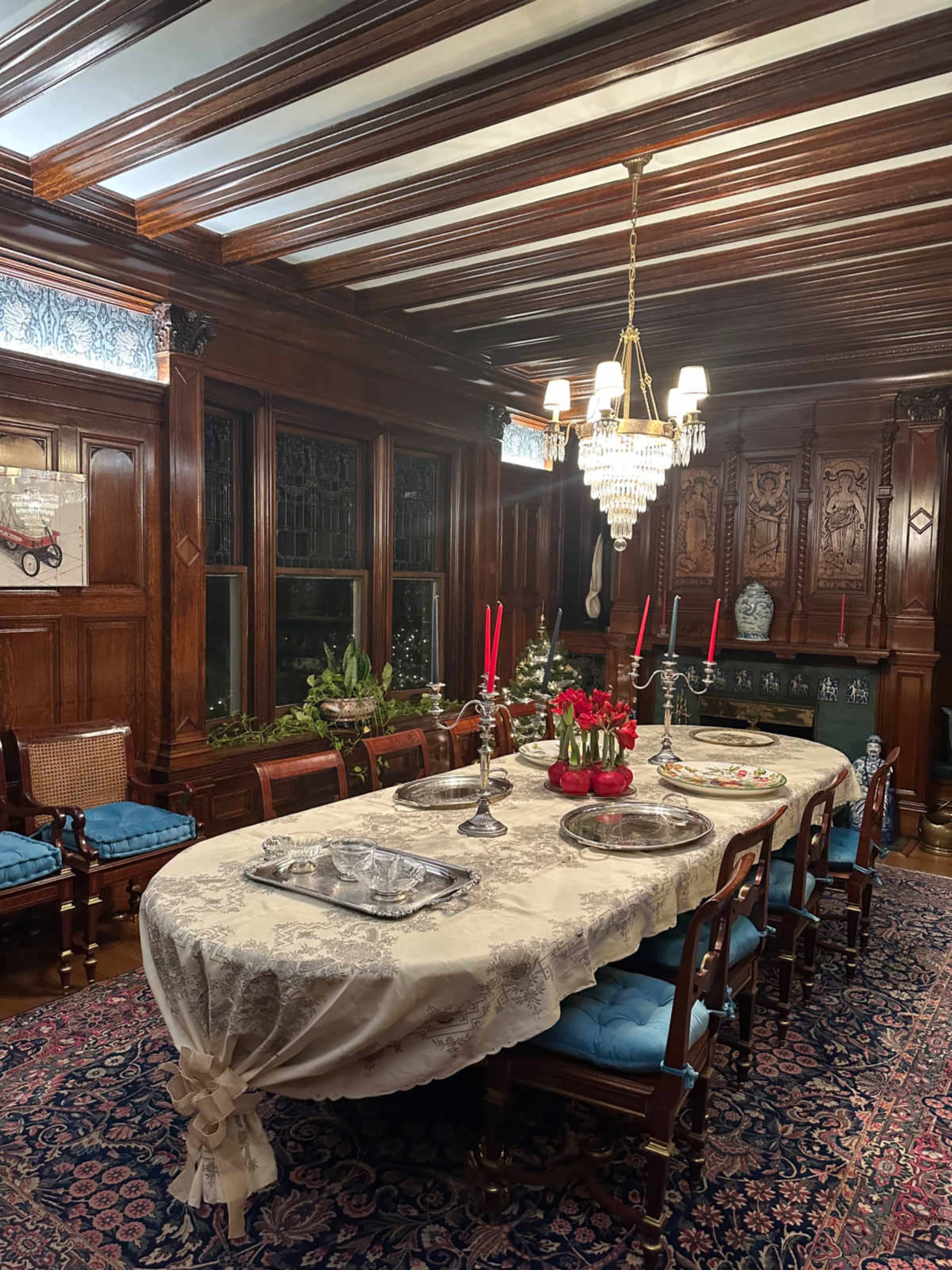 The image shows an elegantly set dining room with a large table covered by a lace tablecloth, surrounded by wooden chairs and decorated with red candles and flowers, all under ornate wooden beams and a chandelier.