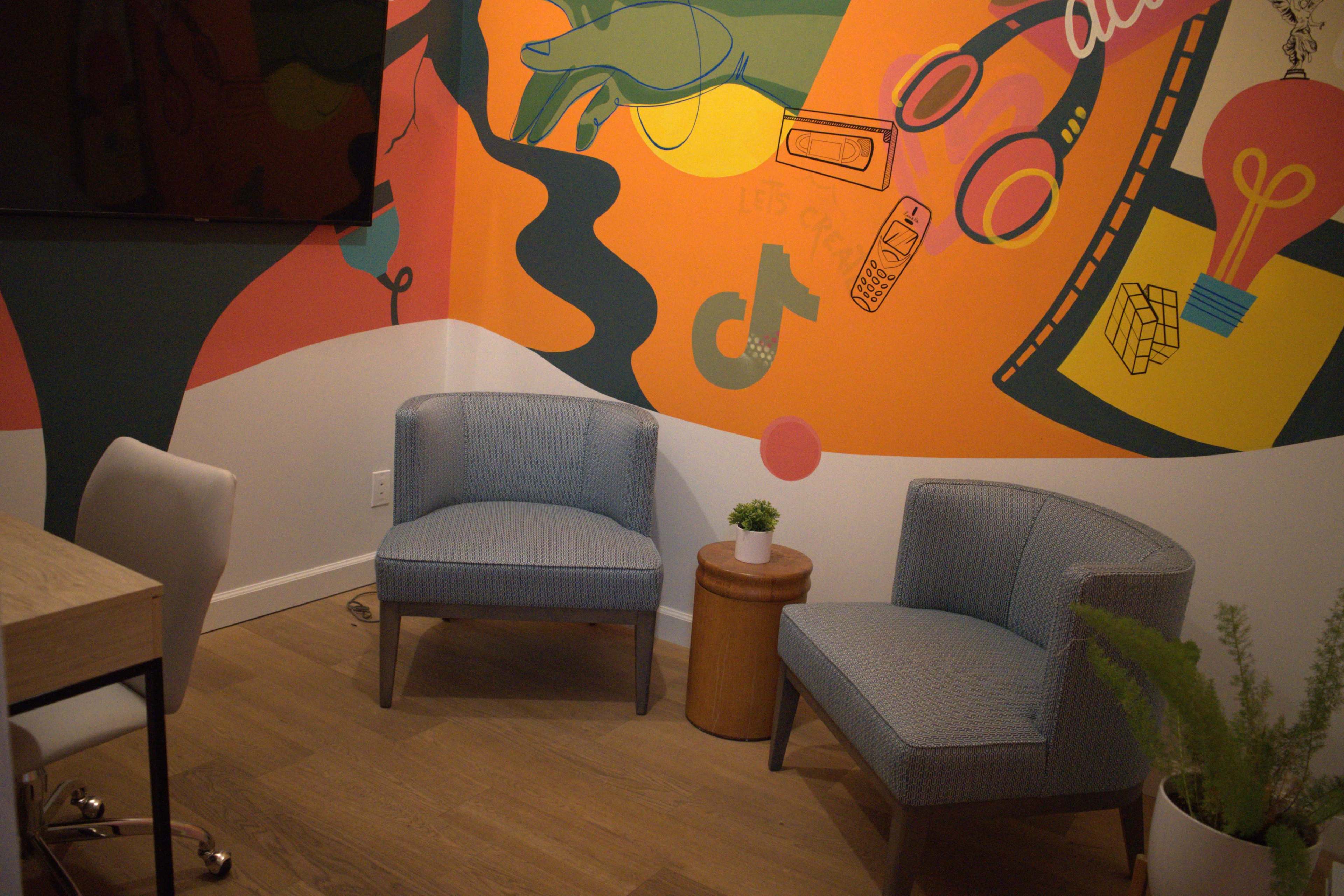 The image shows a colorful mural on the wall of a room with two blue chairs and a small wooden side table.