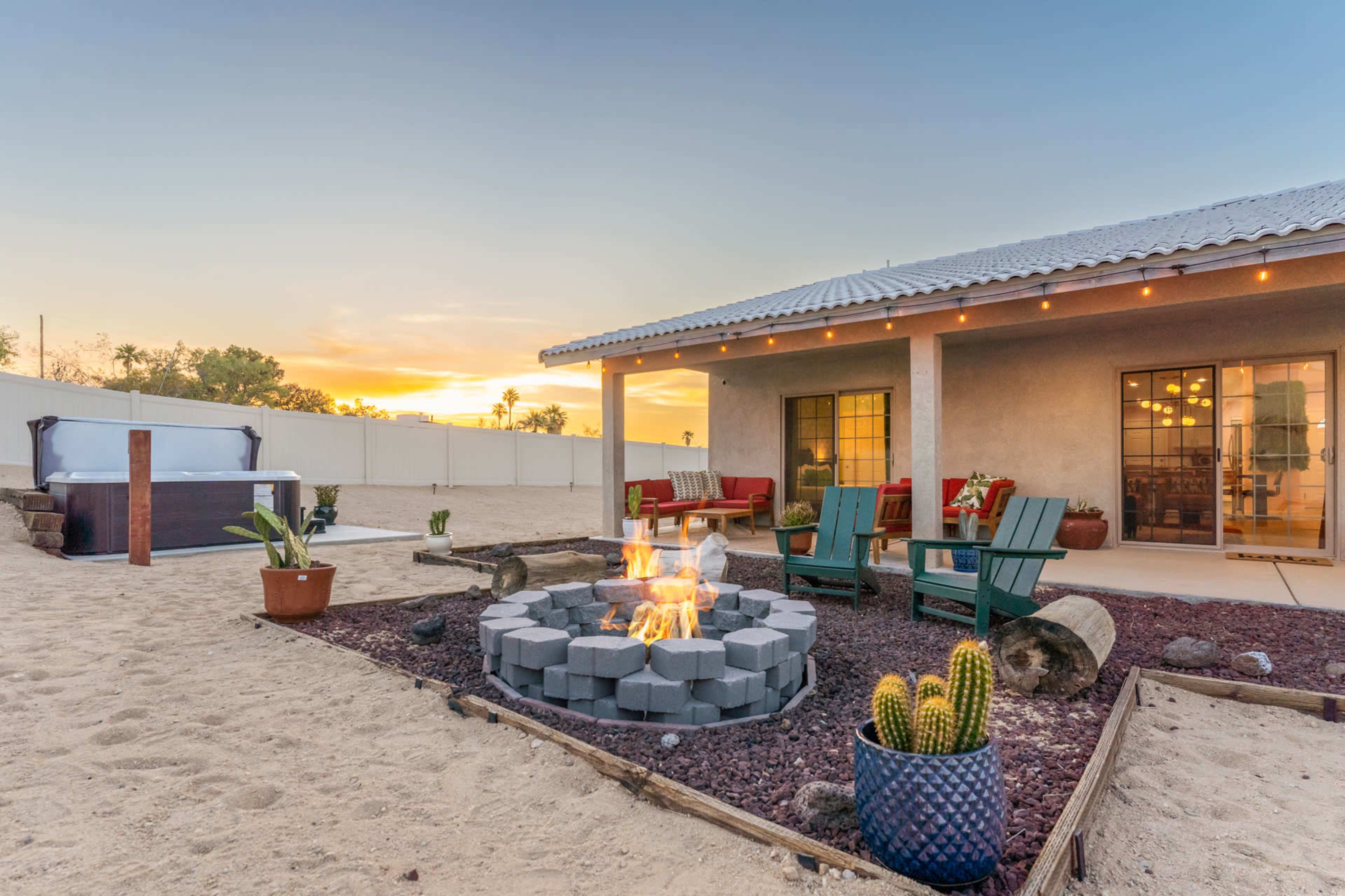 A fire pit surrounded by seating and desert landscaping is illuminated by a sunset in the backyard of a home.