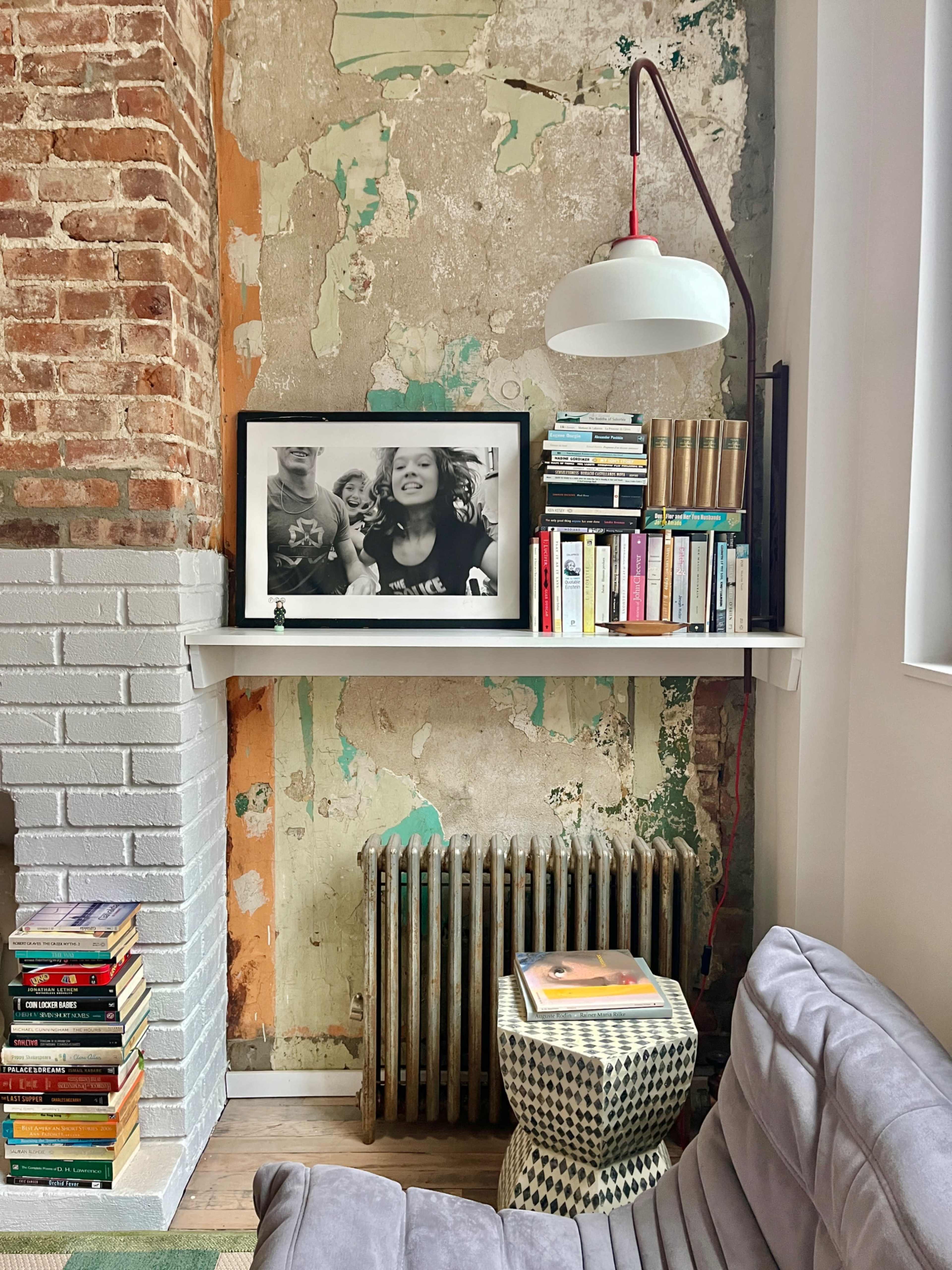 The image shows a cozy living space featuring a shelf with books and a framed black-and-white photo against a weathered brick wall, with a lamp and a radiator nearby.