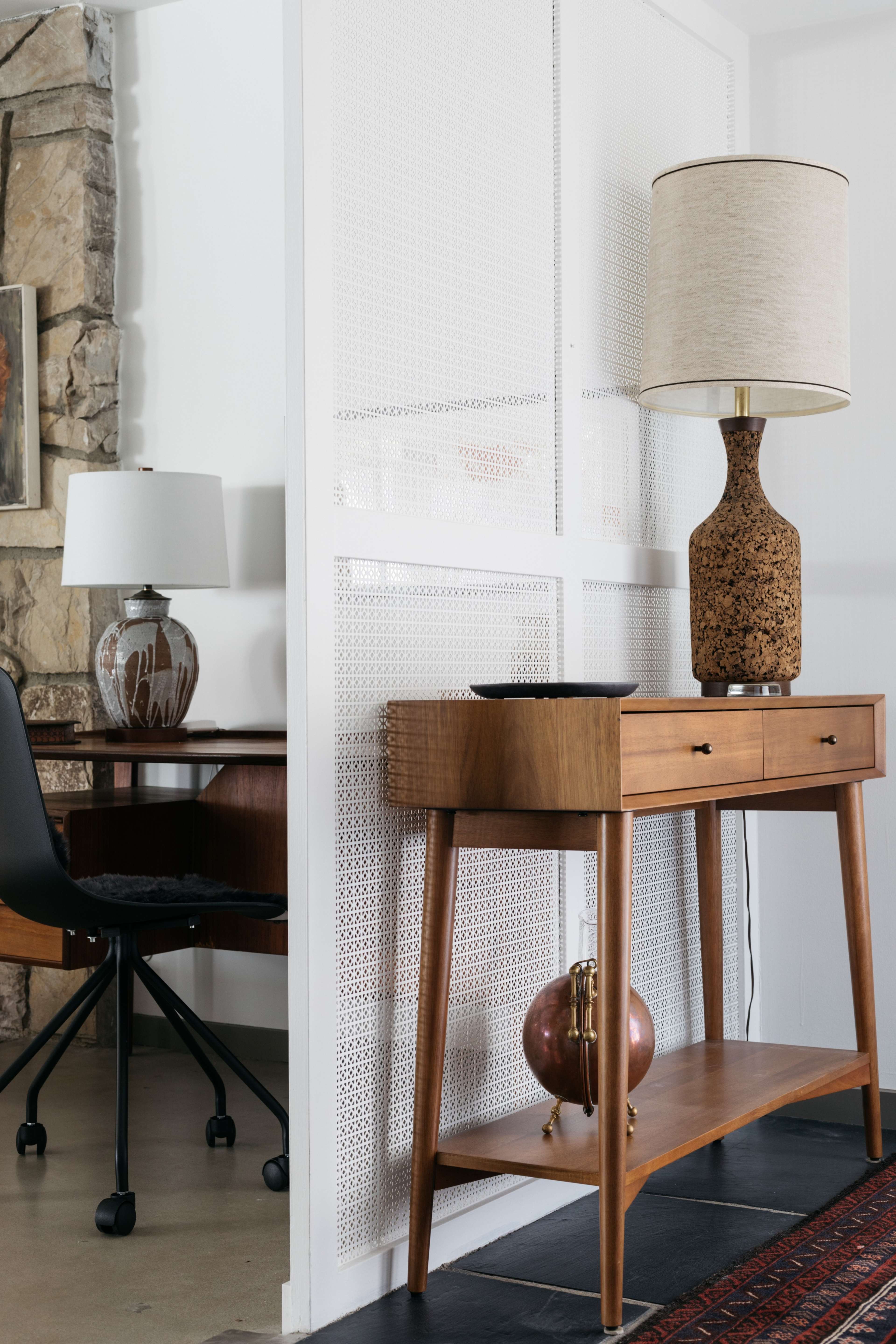 A modern interior setup featuring a wooden console table with a lamp and decorative items, alongside a chair at a desk behind a white perforated panel.