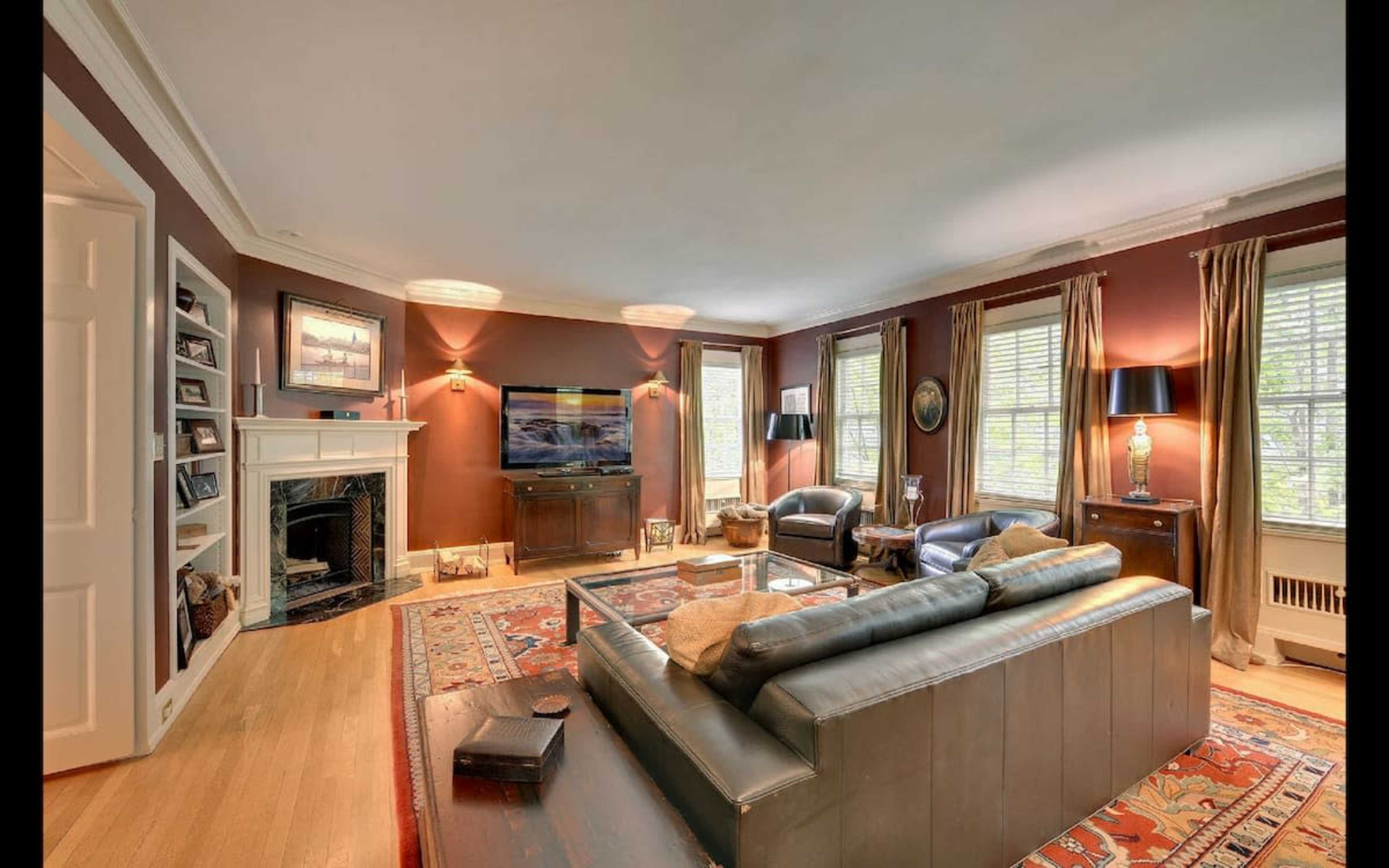 A well-furnished living room with a leather sofa, armchairs, a television mounted on the wall, and large windows allowing natural light.