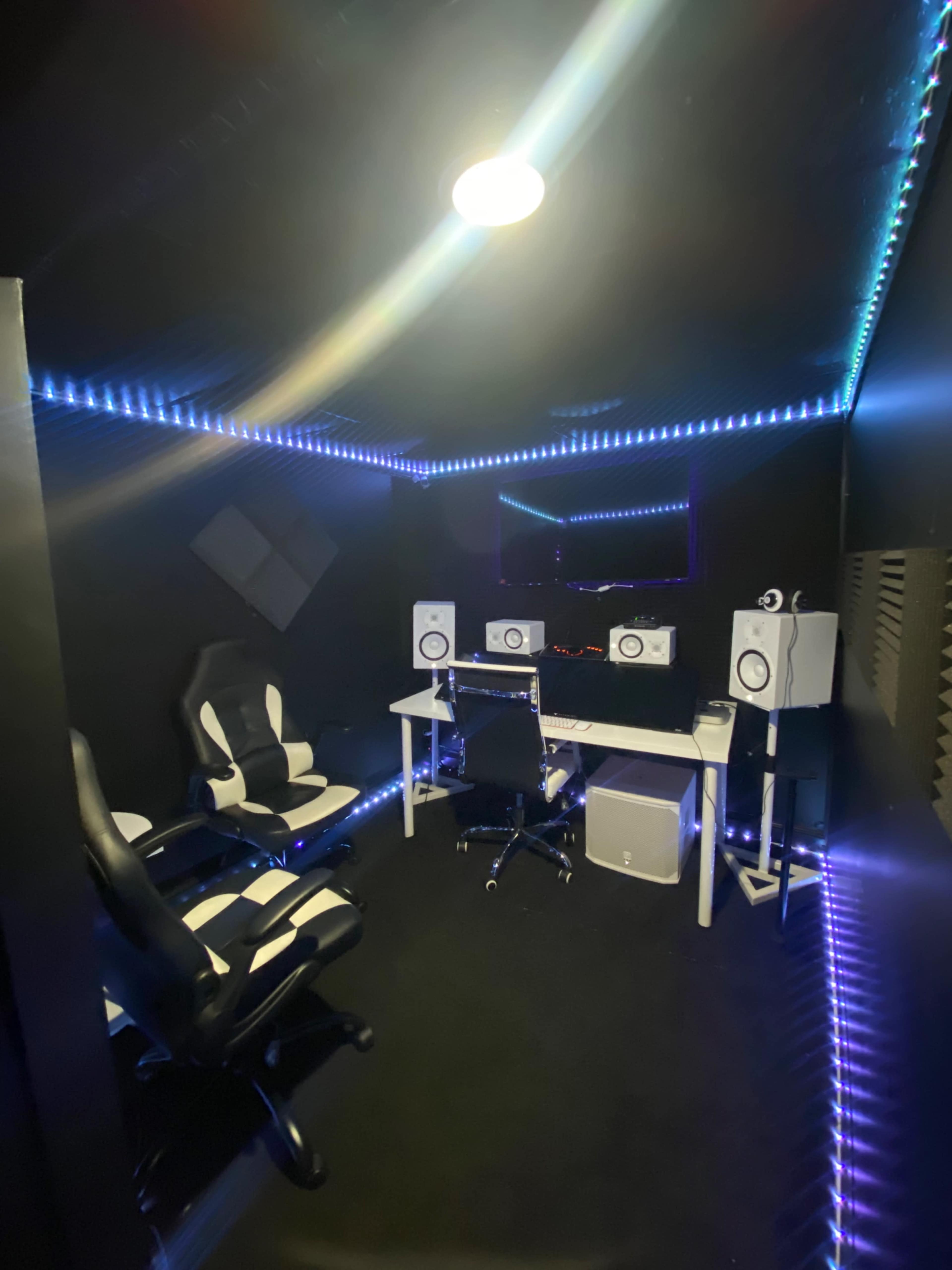 A music recording studio with a black interior, featuring two white monitors, a desk with equipment, and two modern black and white chairs, all illuminated by LED strip lights.
