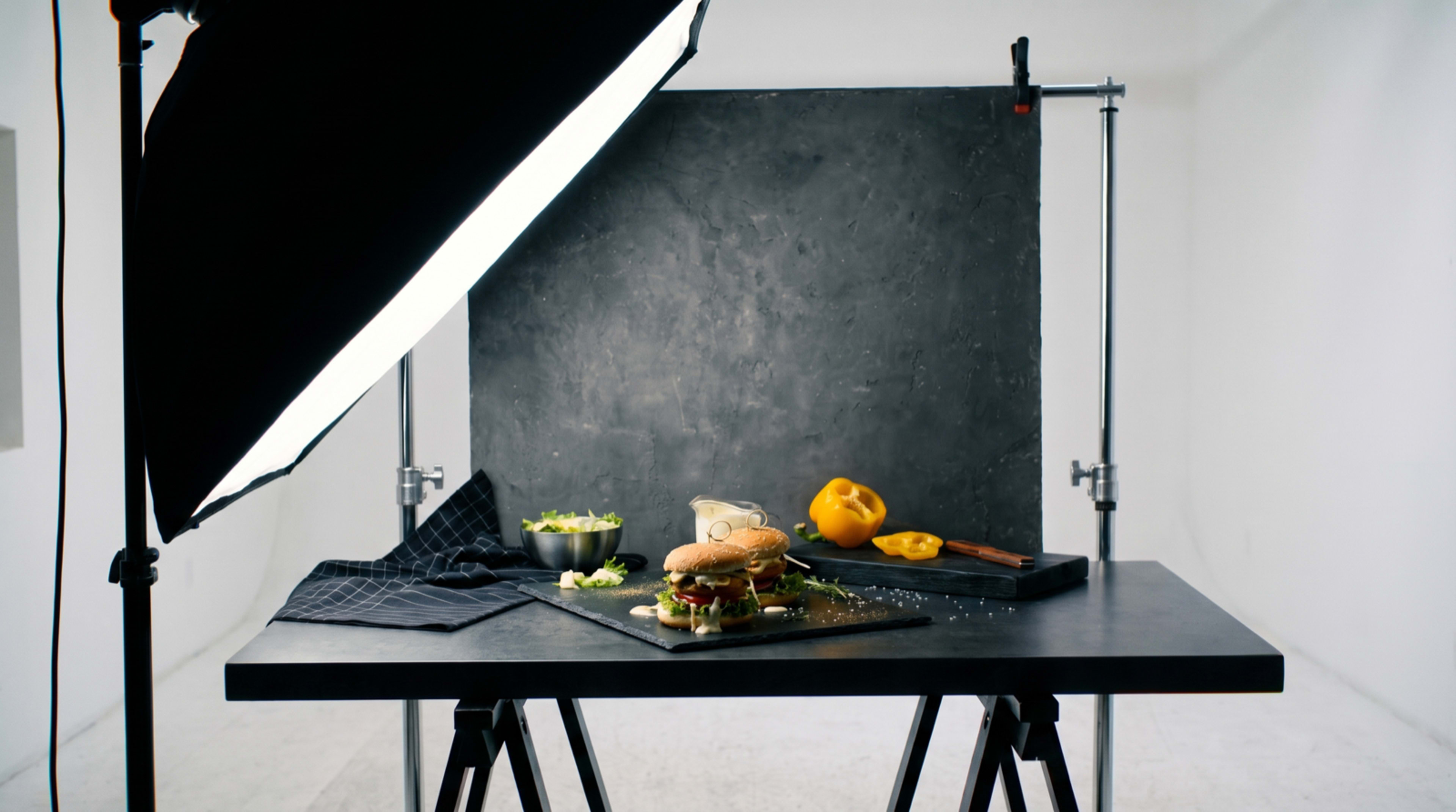 A black table is set up with a burger on a slate platter, accompanied by salad and a chopped yellow pepper, all under bright studio lighting against a gray backdrop.
