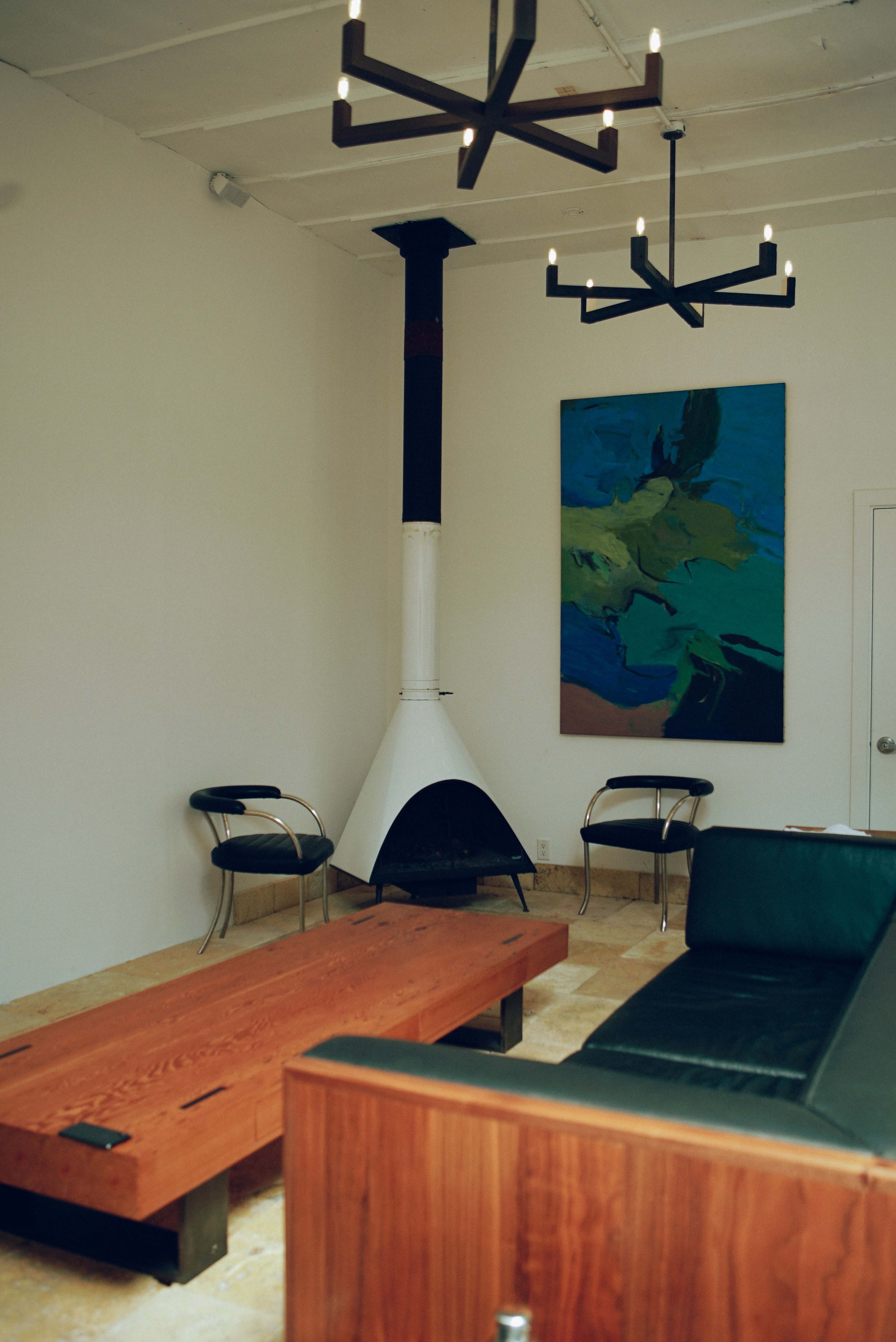 A modern living room features a black sofa, a wooden coffee table, two chairs, a tall black and white fireplace, and an abstract painting on the wall.