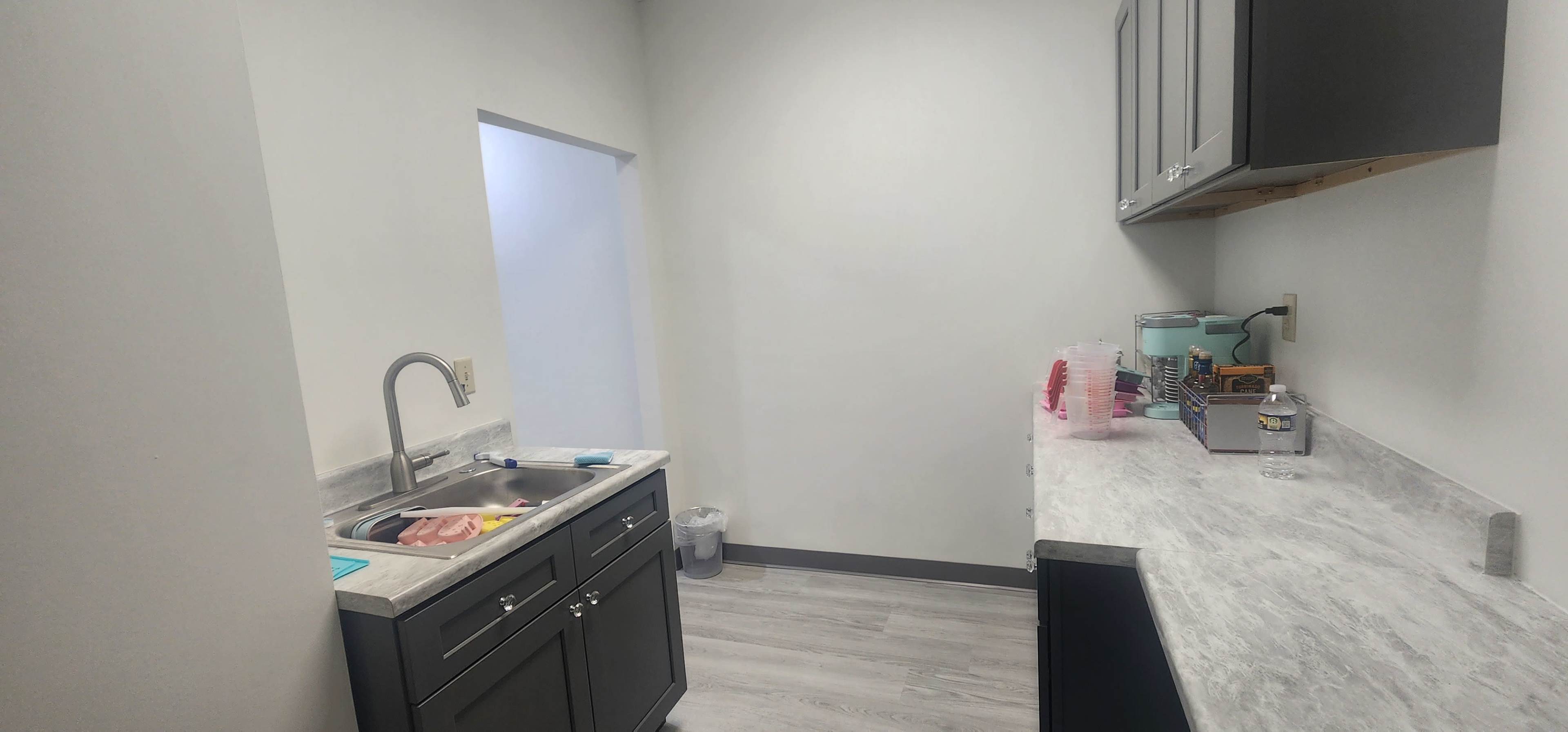 A kitchen area features a sink and counter space with cabinets, alongside a small table displaying utensils and containers.