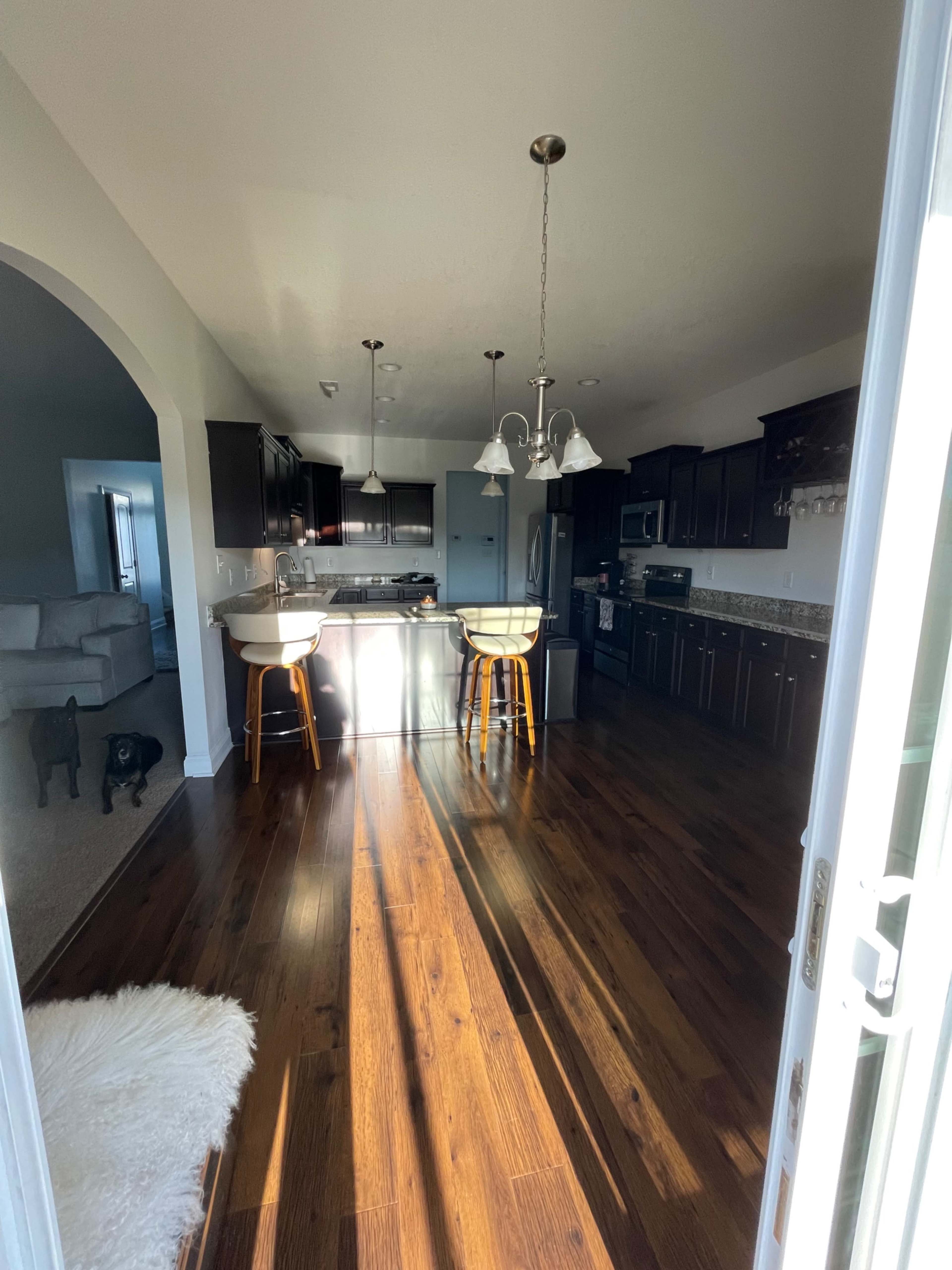 A spacious kitchen features dark cabinetry, a central island with seating, and hardwood floors, illuminated by natural light from the doorway.