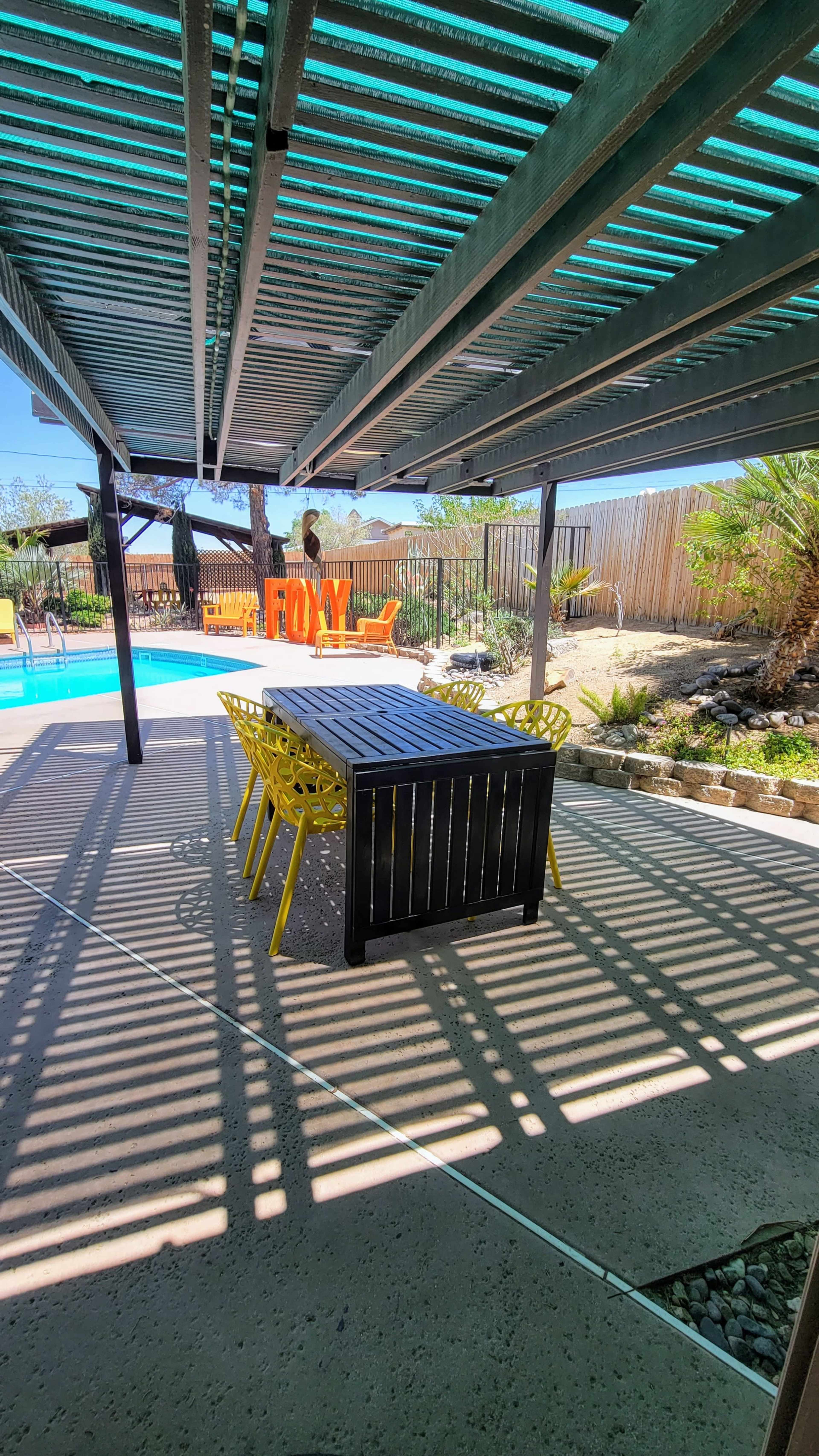 A shaded outdoor dining area features a long black table surrounded by yellow chairs, with a swimming pool and landscaped yard visible in the background.