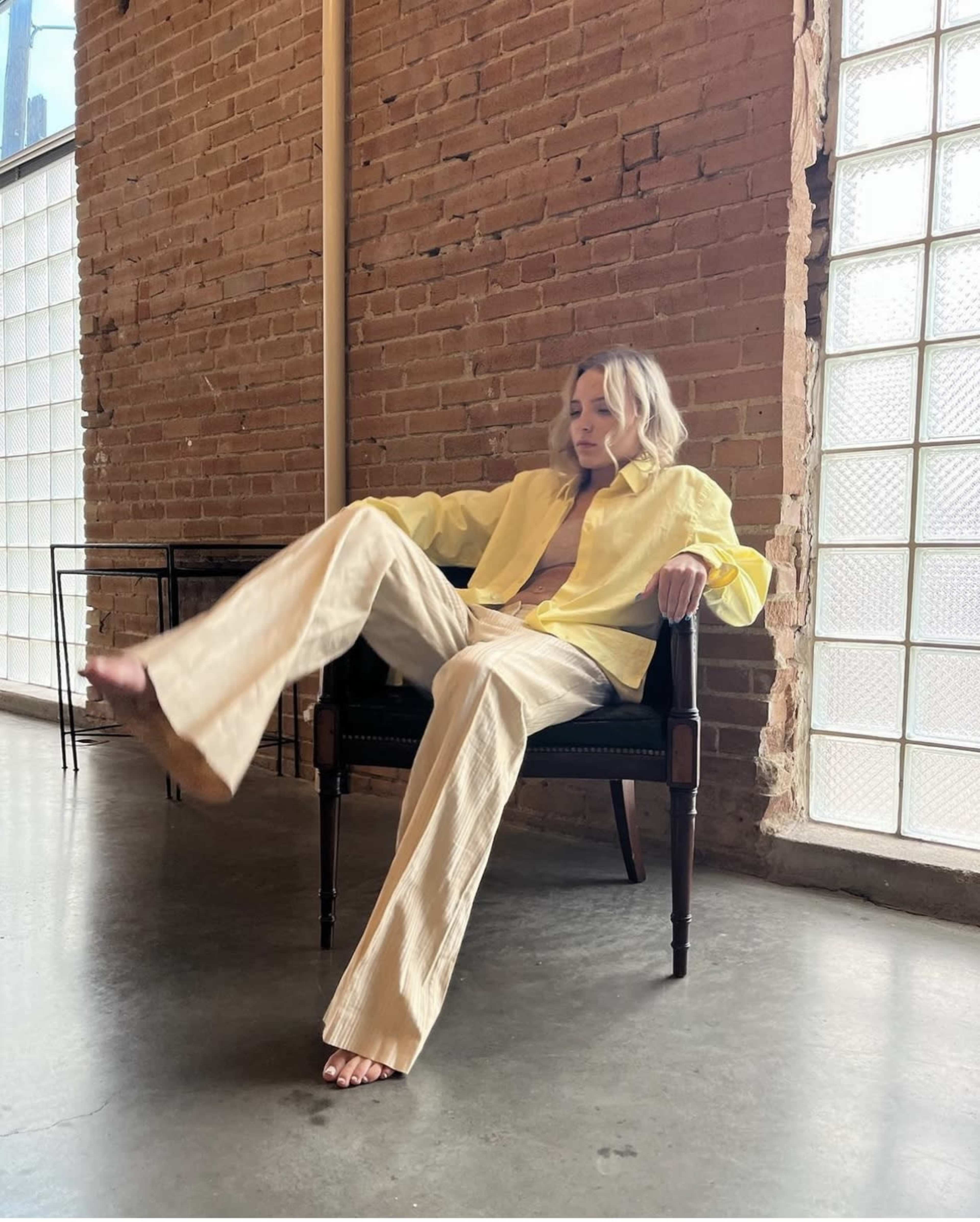 A person with light-colored hair is seated in a chair, wearing a yellow shirt and beige flared pants, in a room with brick walls and large windows.