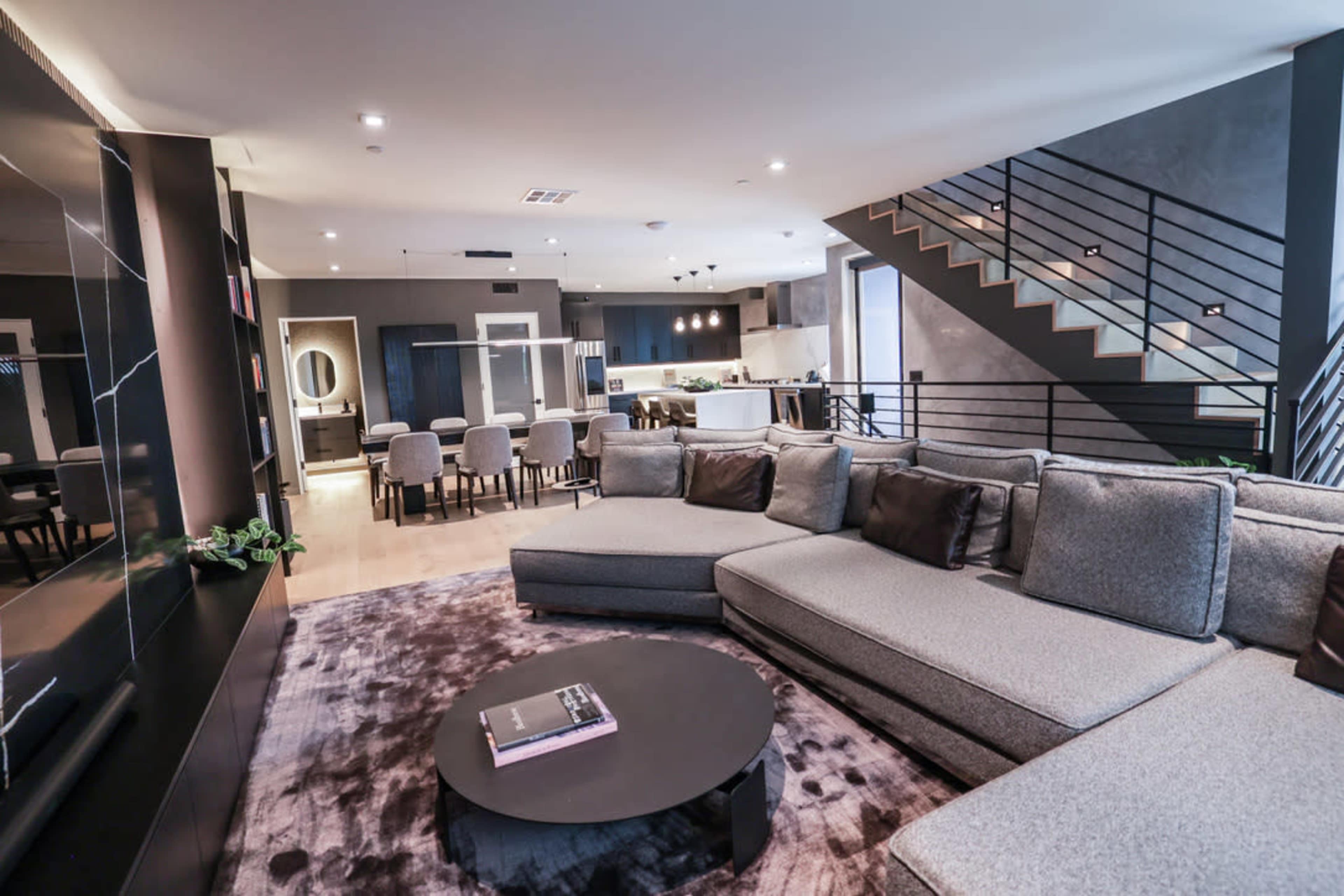 A modern living room with a gray sectional sofa, a round coffee table, and an open layout leading to a dining area and kitchen.
