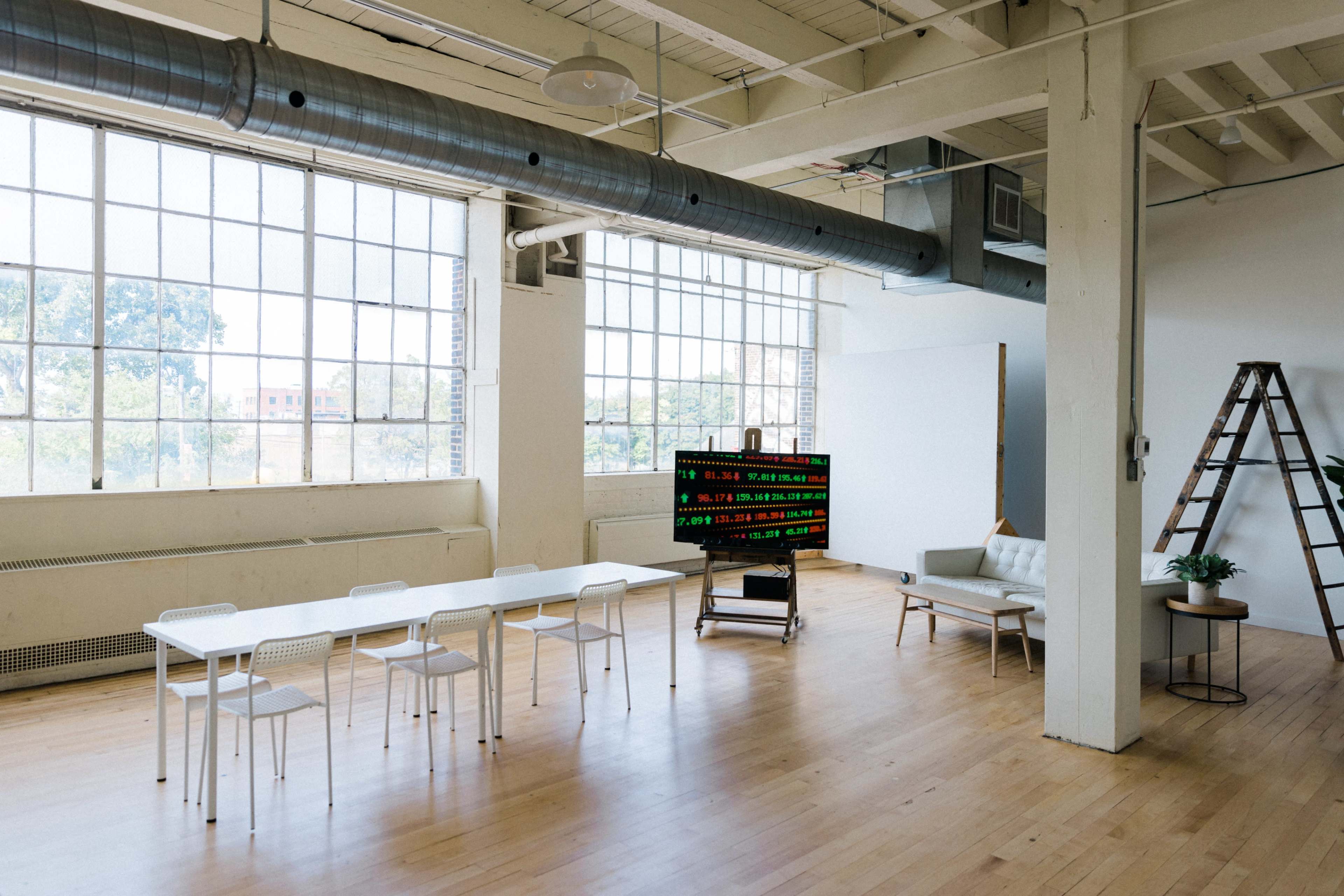 A spacious, well-lit interior featuring a long table with white chairs, a couch, a large display monitor showing data, and a ladder against the wall.