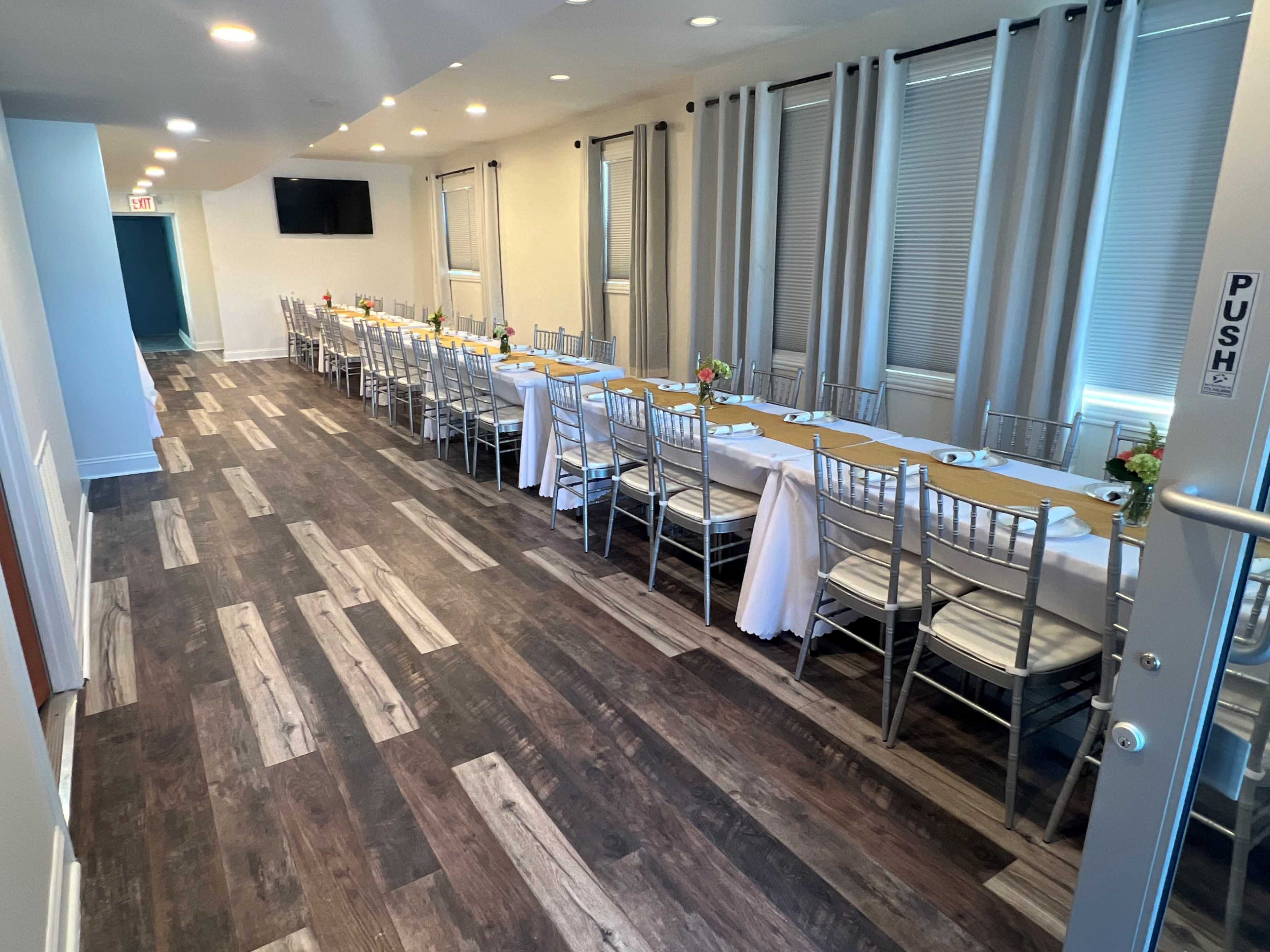 A long dining table is set with white tablecloths and chairs in an elegantly arranged hallway with light-colored walls and wood flooring.