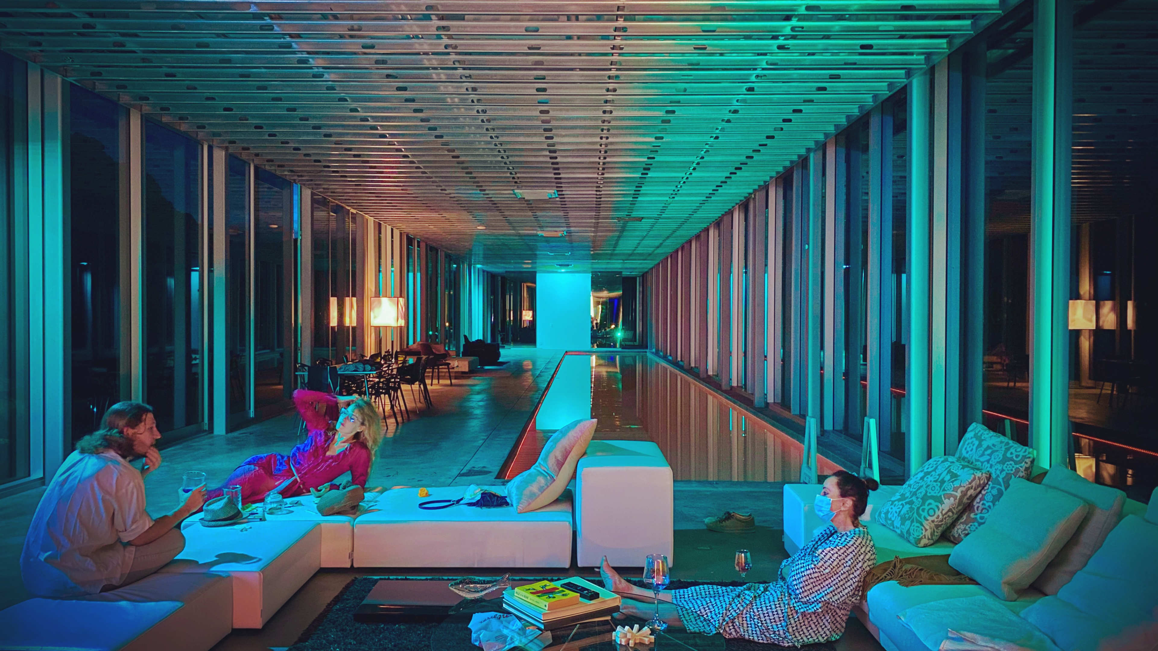 The image shows a modern, brightly lit indoor area with a long pool and several seated individuals lounging on sofas, engaged in conversation.