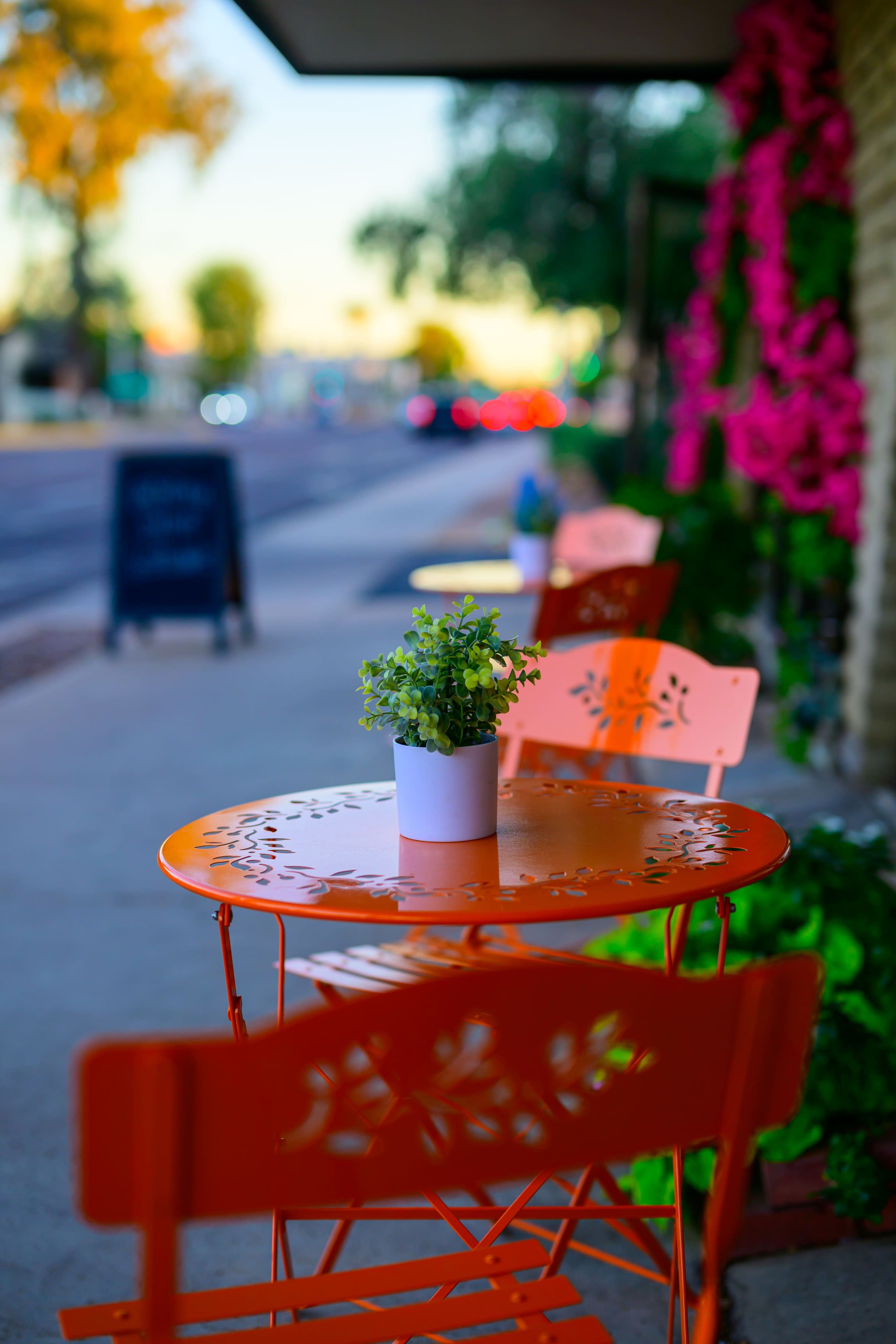 A small orange table with a white potted plant is set up on the sidewalk adjacent to a street lined with colorful flowers.