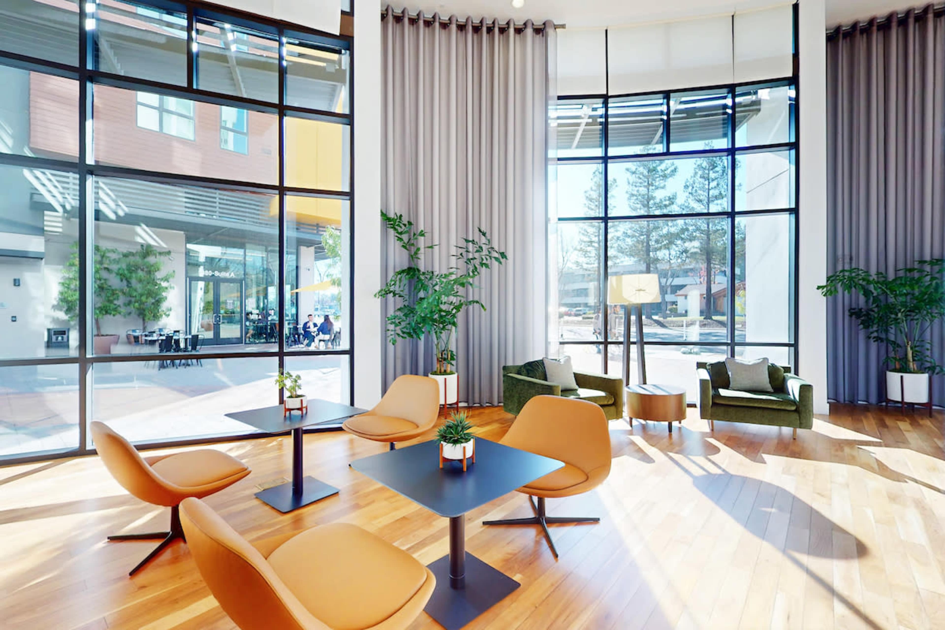The image shows a modern interior space with large windows, featuring wooden flooring, several orange chairs, black tables, and potted plants.