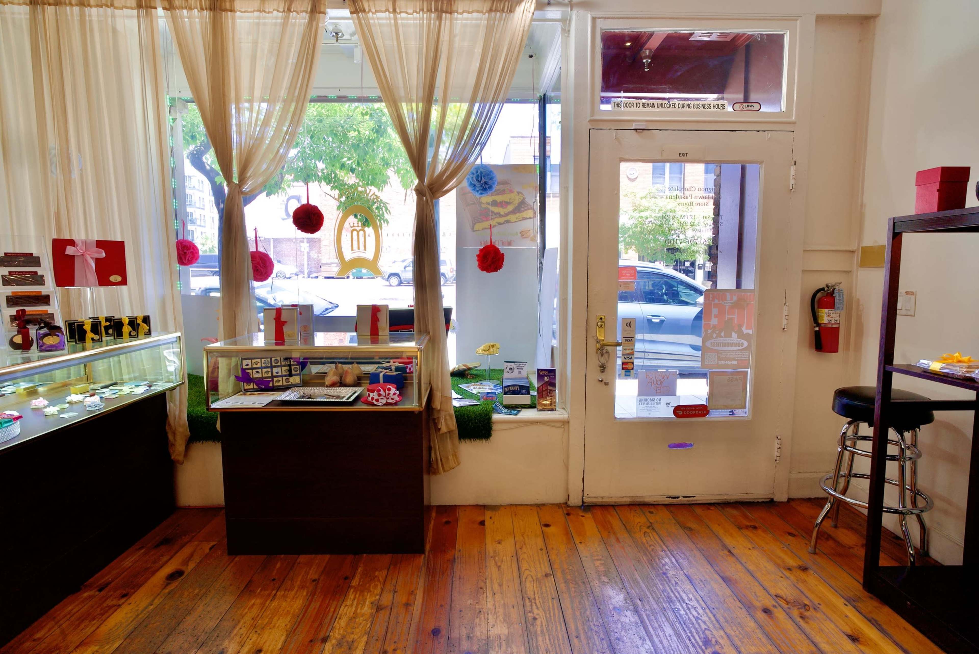 The image shows a brightly lit storefront with a wooden floor, display cases filled with colorful items, and large curtains framing the entrance.