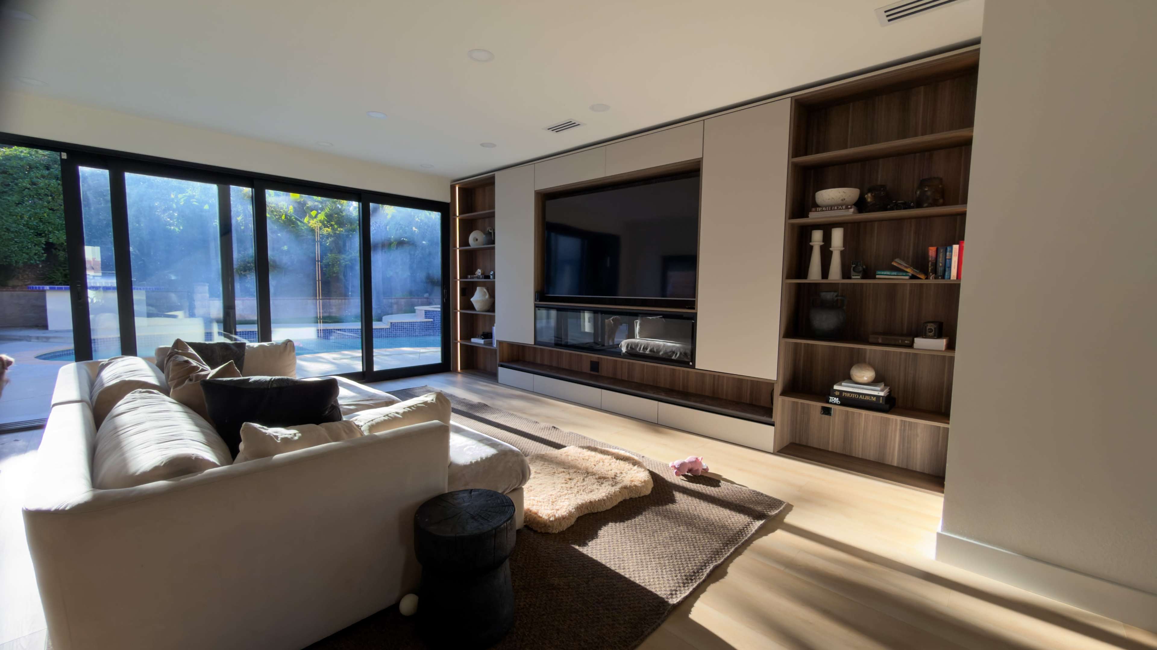 The image shows a modern living room with a large wall-mounted TV, built-in shelving, a beige sofa, and a view of an outdoor space through expansive glass doors.