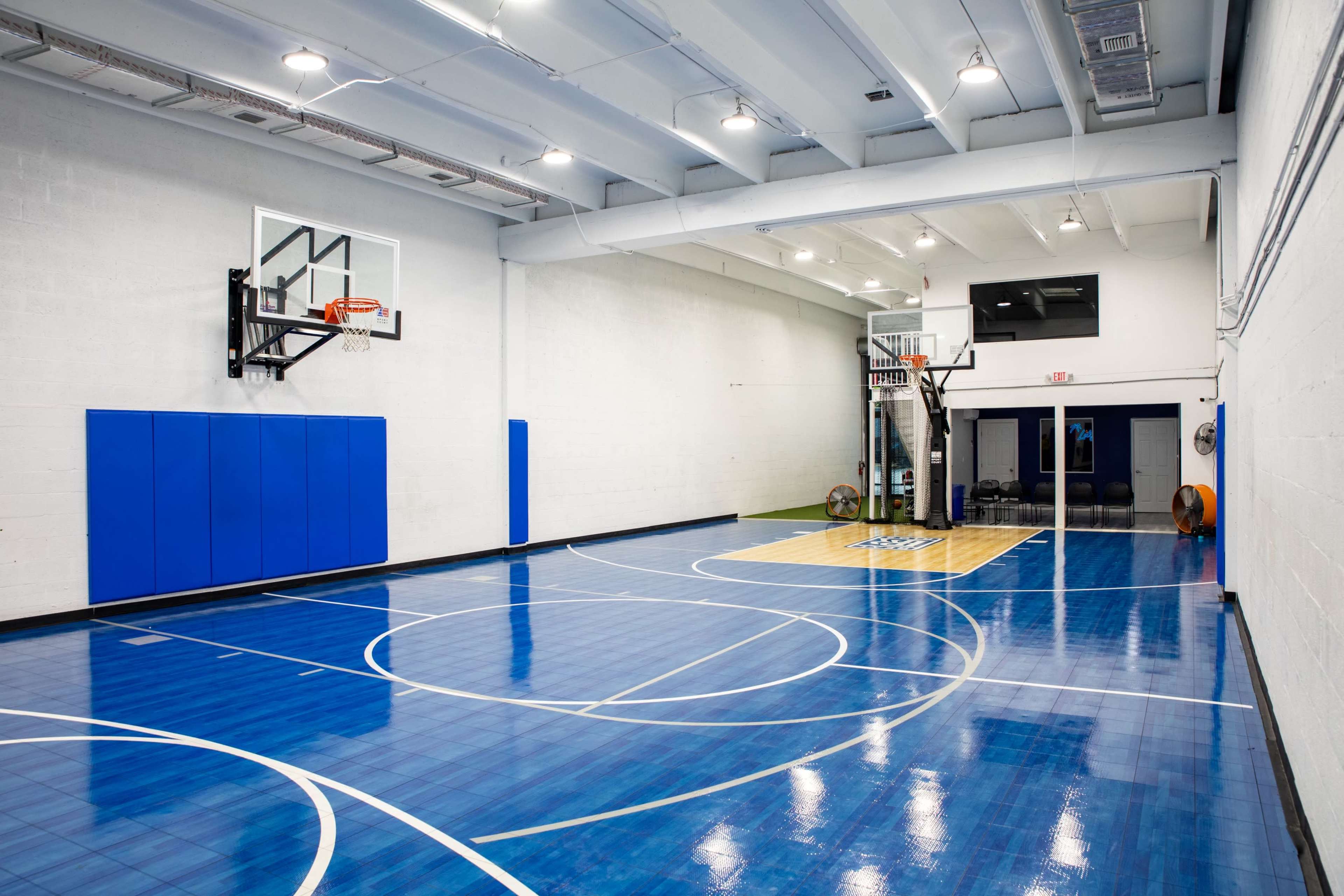 A basketball court with a blue and yellow floor, a hoop mounted on the wall, and padded wall sections.