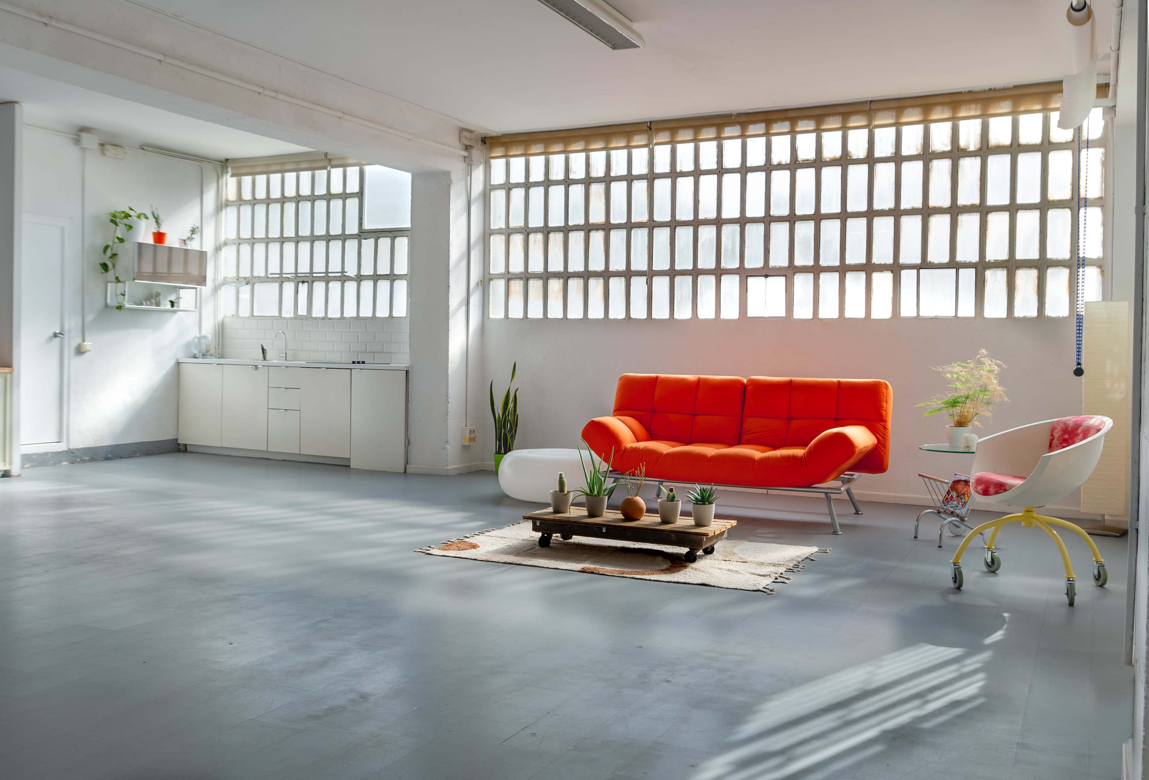 A bright, open living space features an orange couch, a wooden coffee table with decorative items, and a small kitchen area in the background.